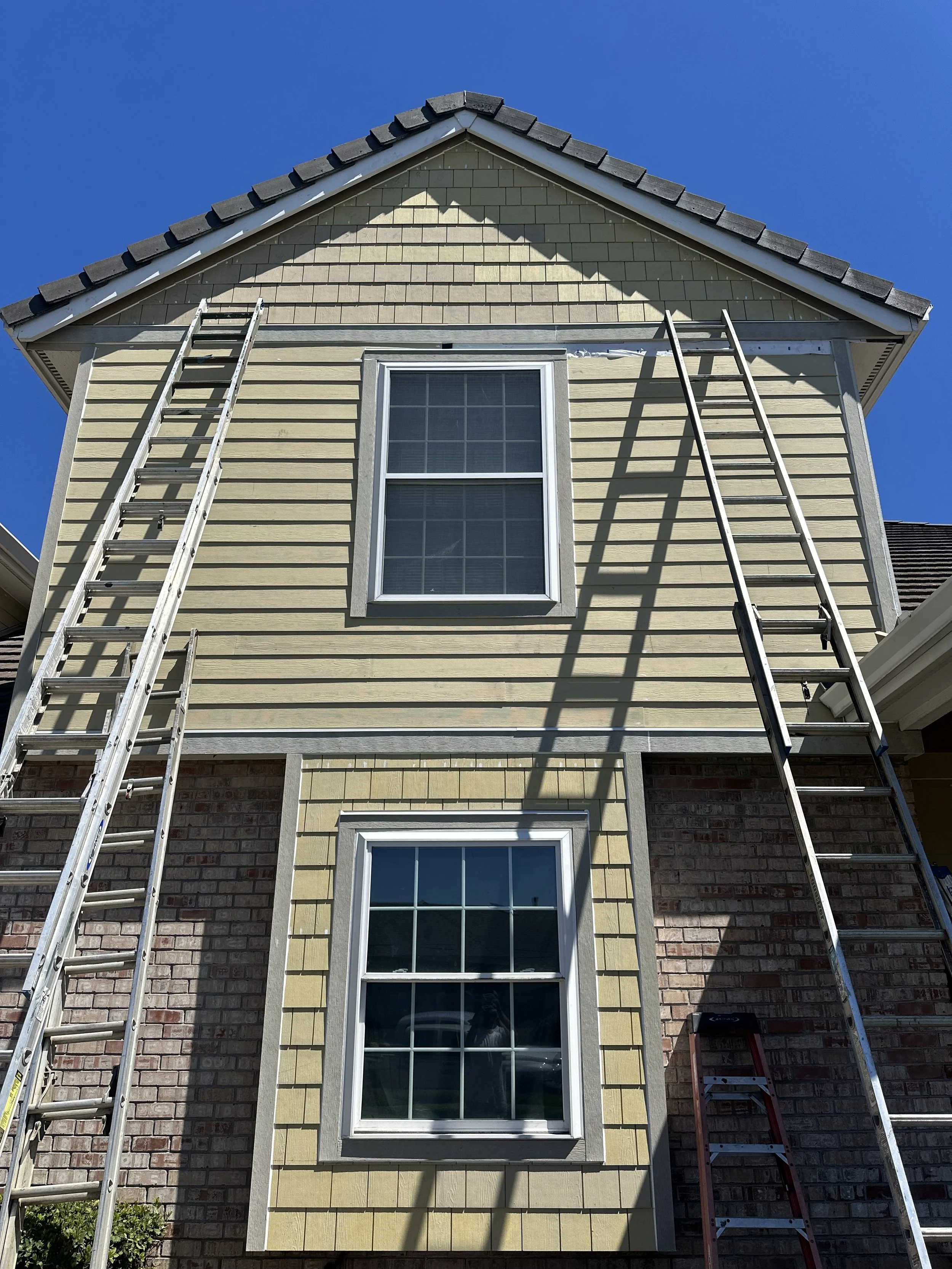 Two ladders leaning against the exterior of a house with beige siding, a window, and an upper section with shingles, under a clear blue sky.