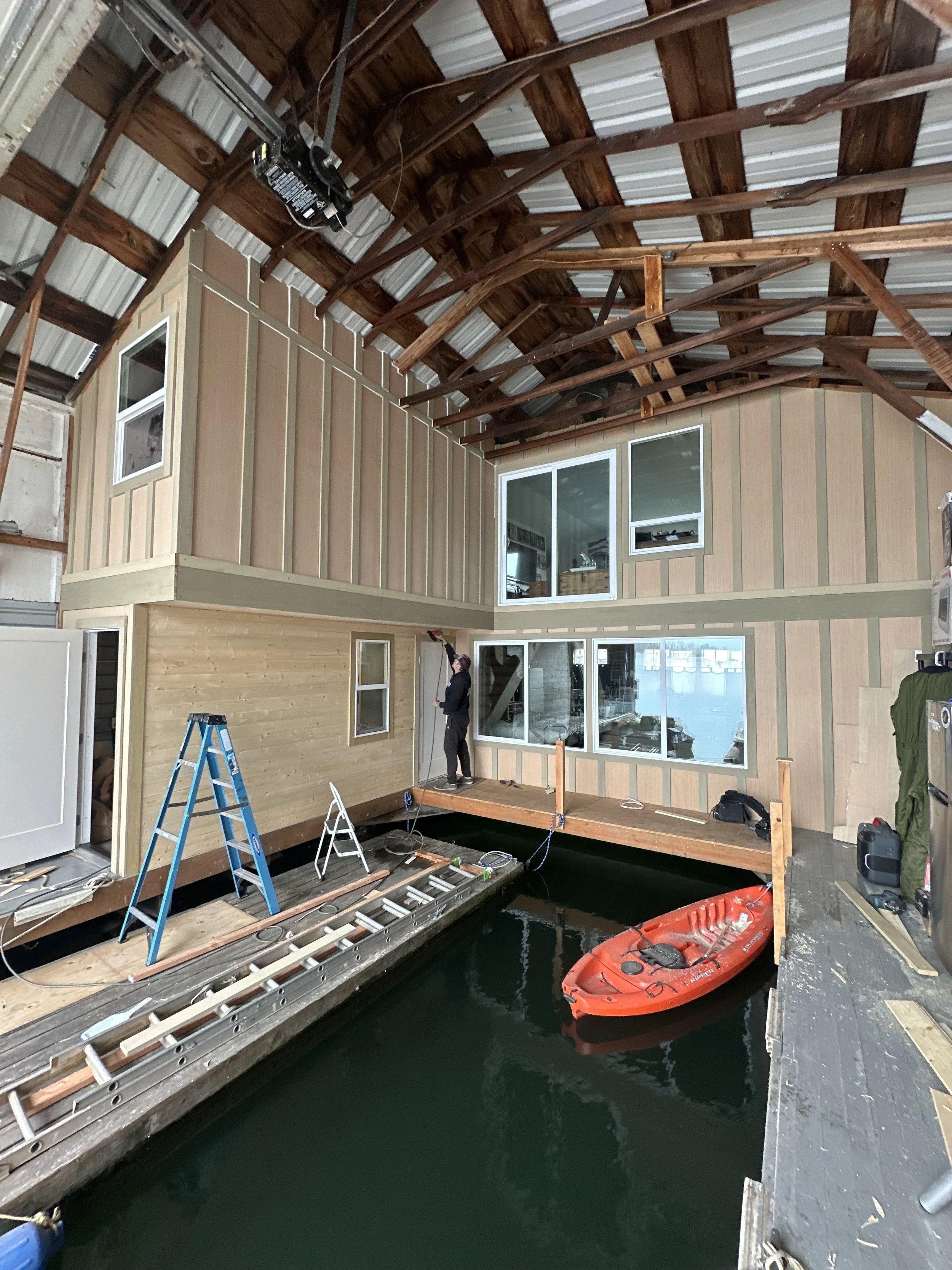 Construction inside a building with unfinished walls and exposed wooden beams. A person in dark clothing is using tools, standing on a wooden platform near water. An orange kayak is floating in the water next to the platform, and ladders and construc