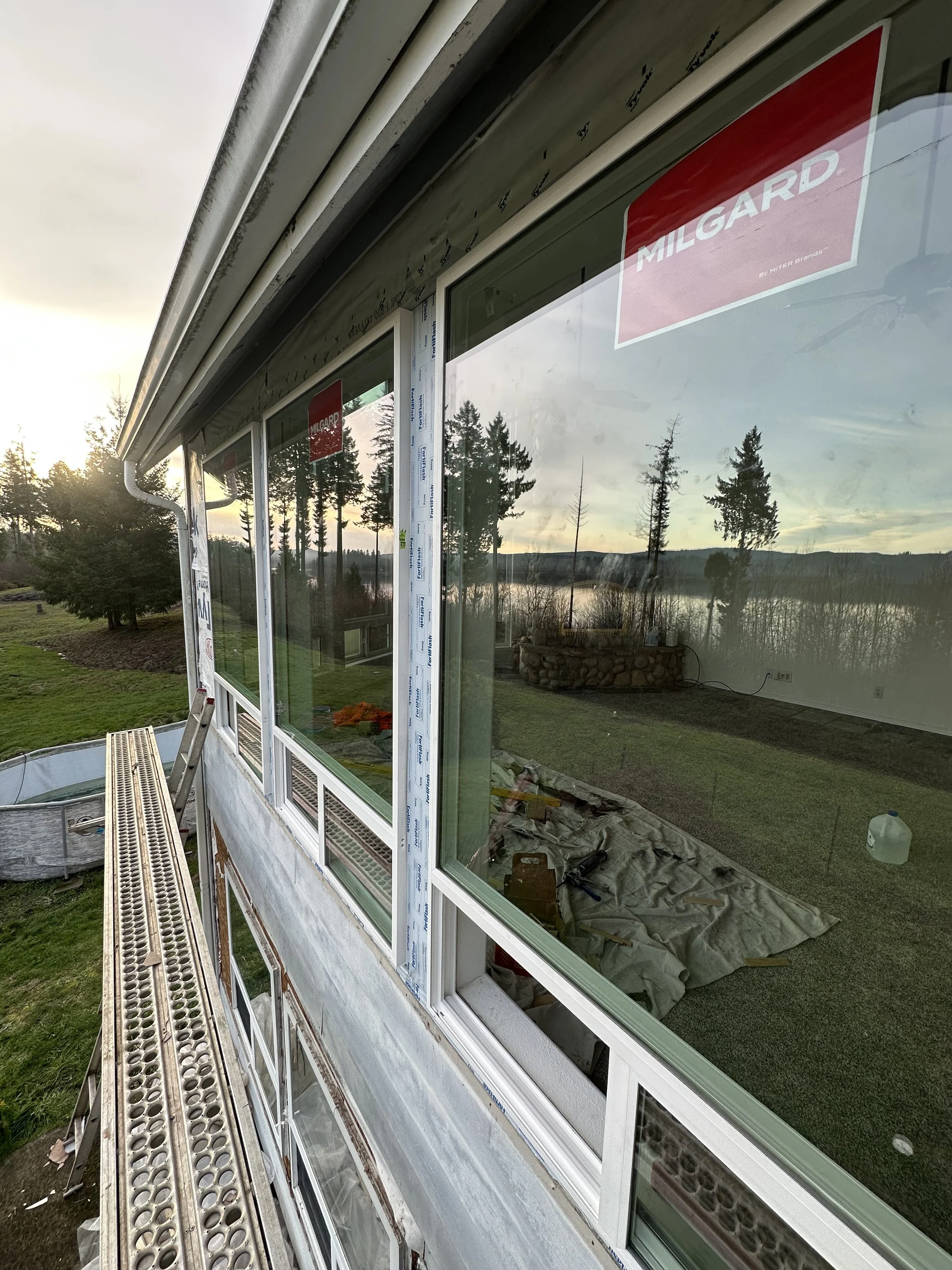 Construction workers installing large glass windows on a building with scenic outdoor view of trees, water, and cloudy sky.