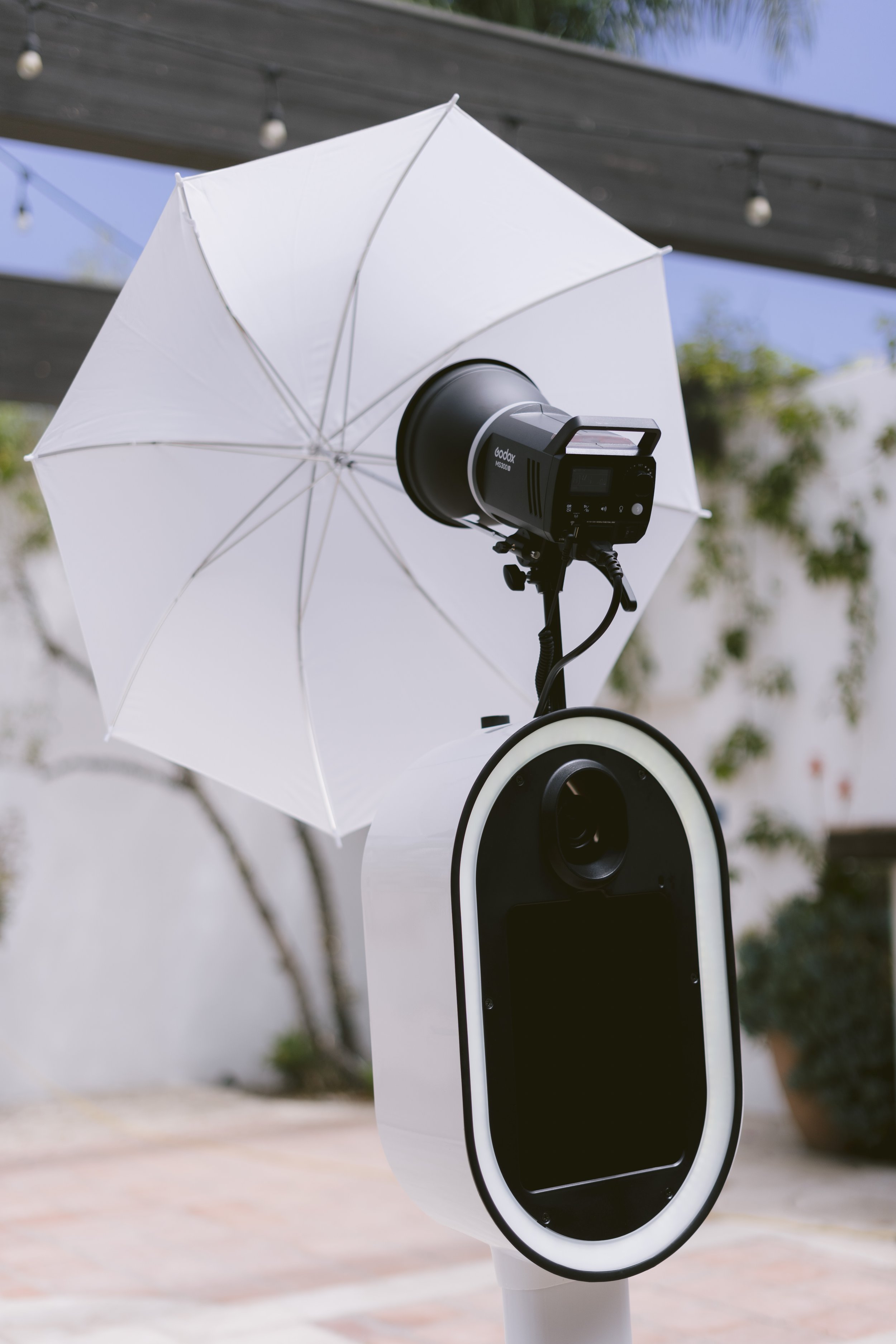 A photo of an automatic contactless photo booth, which includes a black and gray camera mounted on a white stand with a circular frame, a white umbrella for lighting, and a black card reader or scanner attached to the stand, set outdoors with blurred trees and sky in the background.