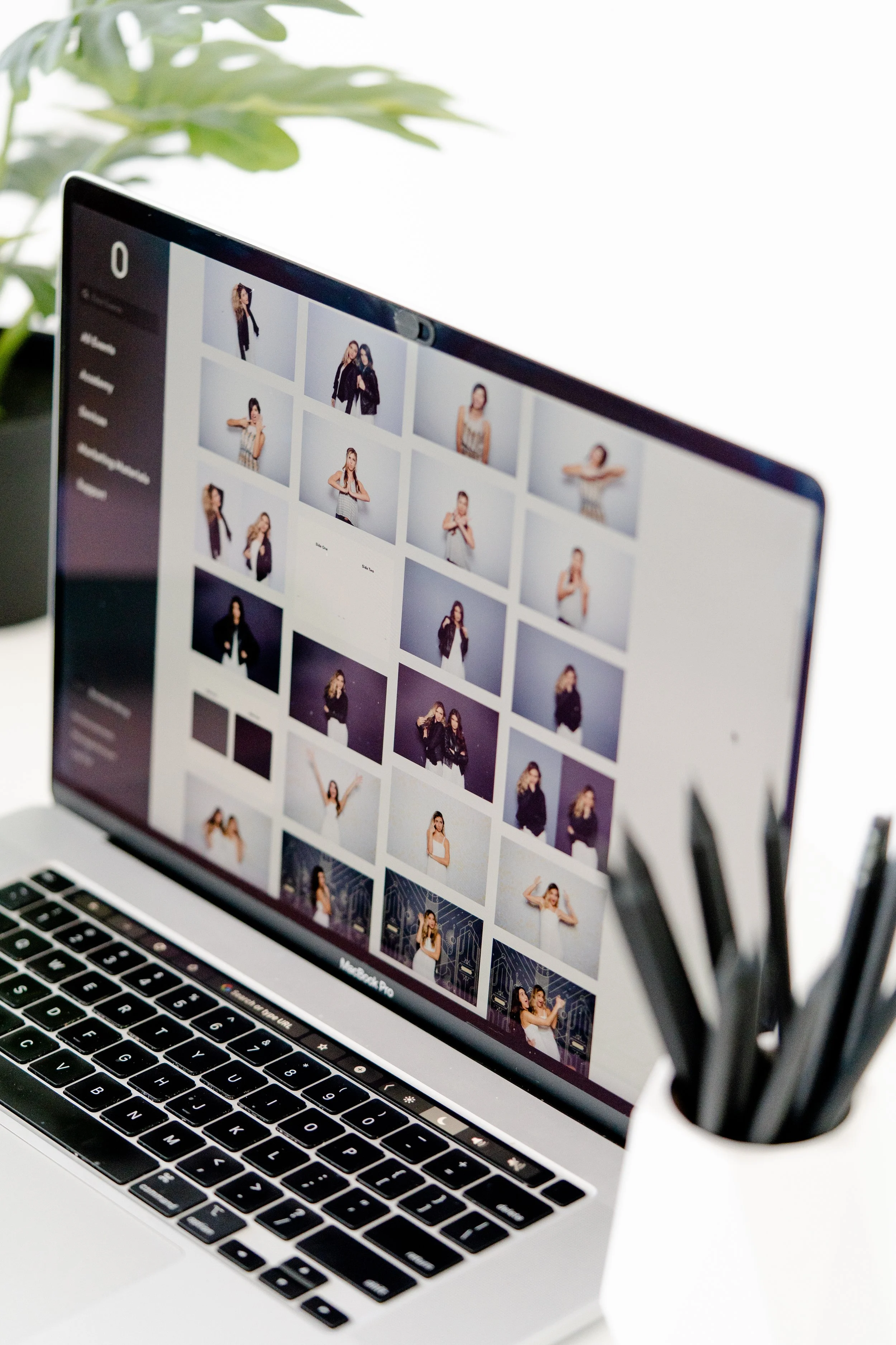A MacBook Pro laptop displaying a photo editing software with a grid of portrait images of women on the screen, placed on a white desk with a plant in the background.