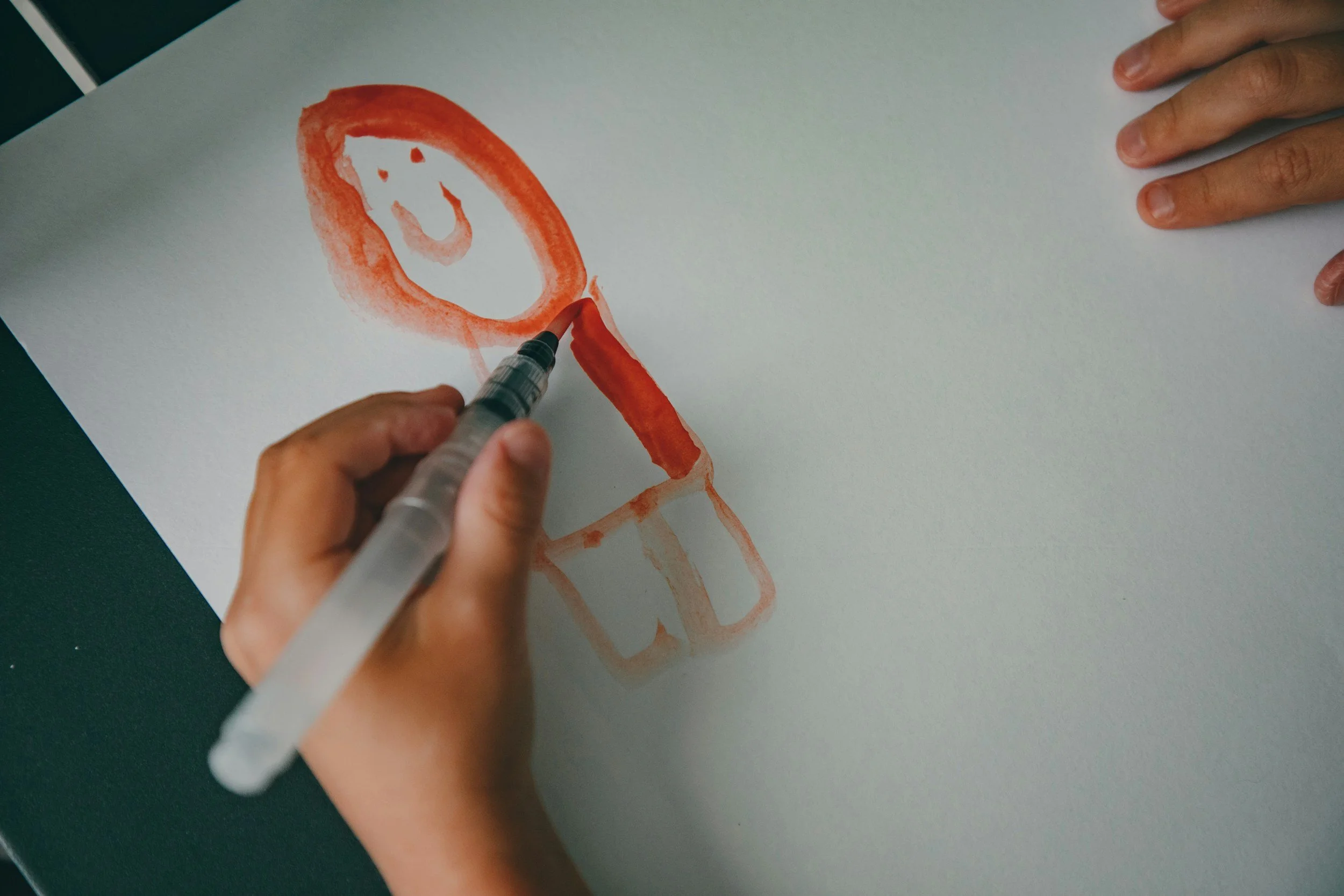 A person is drawing or painting a portrait of a woman on a white sheet of paper using a red-orange marker, with a hand supporting the paper on the right side.