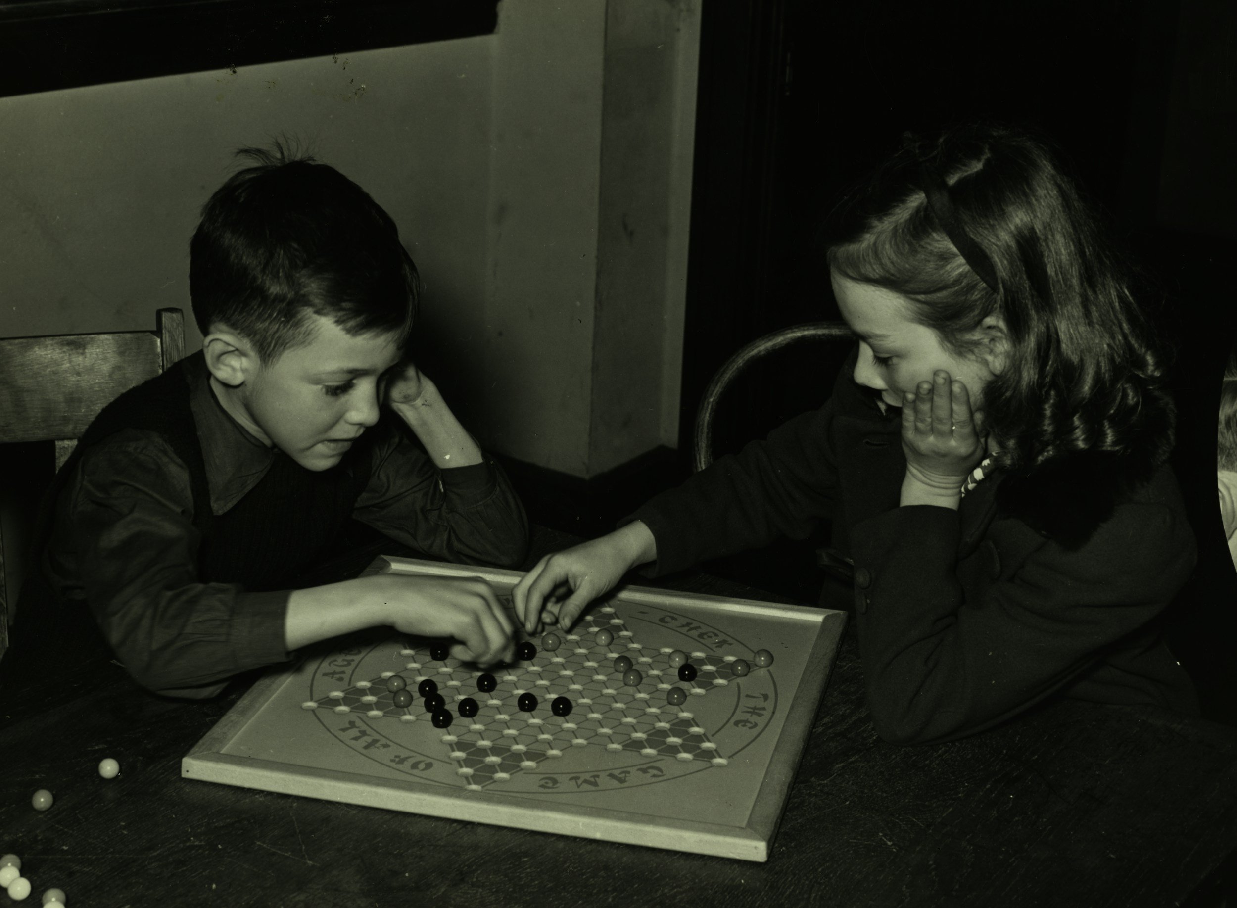 Two children playing the game of Go at a table, with black and white stones. The boy on the left has dark hair and is wearing a dark jacket. The girl on the right has wavy hair with a headband and is resting her chin on her hand, gazing at the game.
