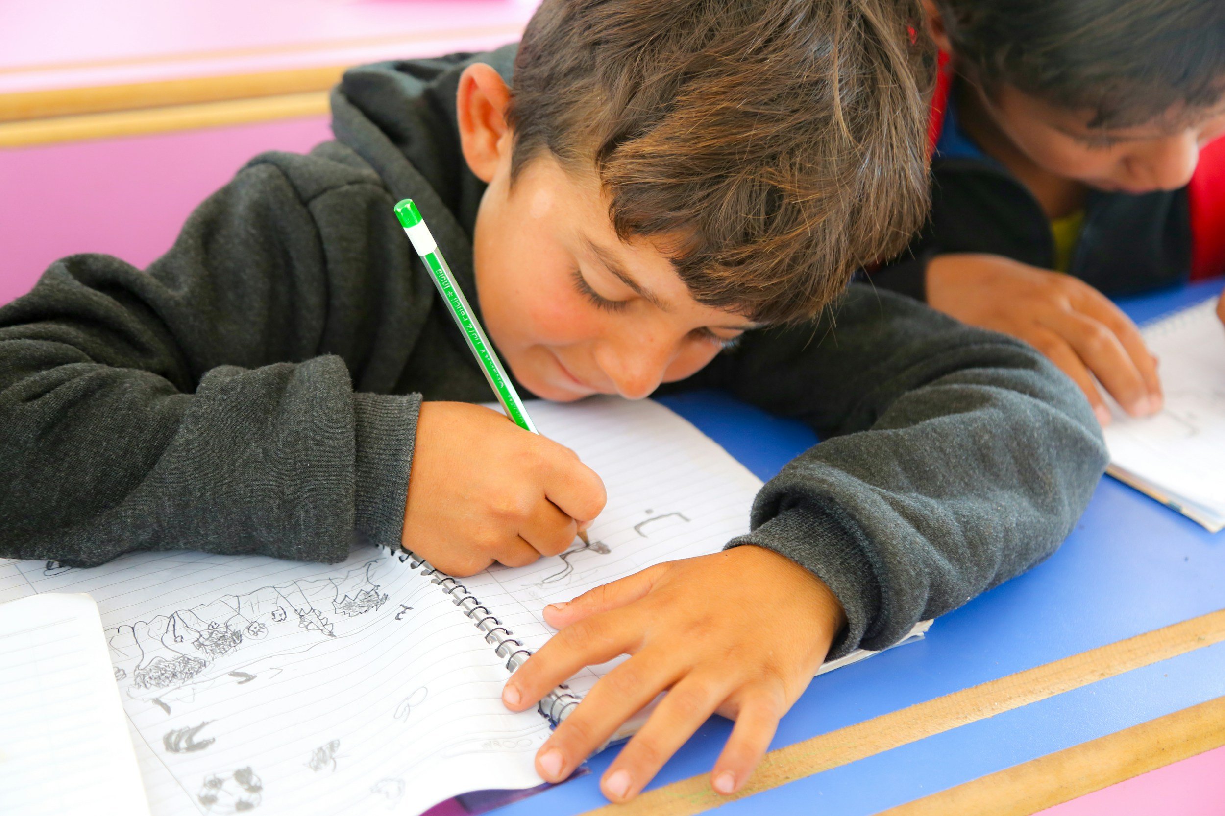 A young boy with brown hair and a dark sweatshirt is drawing on a spiral notebook with a green pencil. His head is tilted down, and he is focused on his drawing, which includes various sketches of what appear to be animals or objects. Another child is visible beside him, also engaged in drawing, on a blue surface with a pink and yellow background.