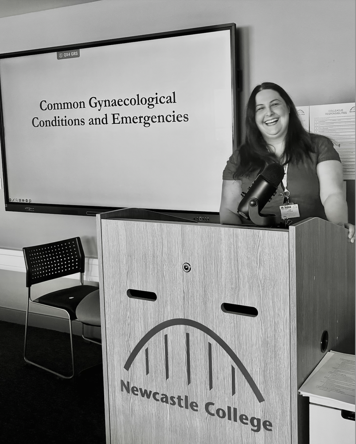 A woman smiling and speaking at a Newcastle College lecture on gynecological conditions, standing behind a wooden podium with a microphone.