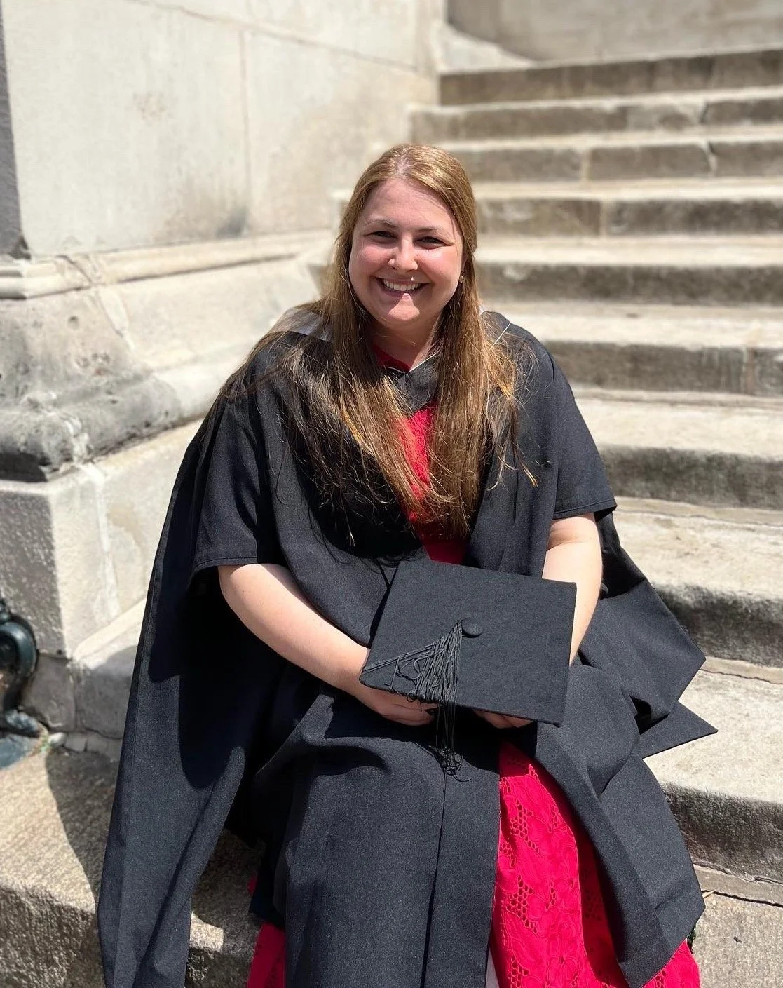 A young woman in a graduation gown sitting on outdoor stone steps, holding a graduation cap, smiling.
