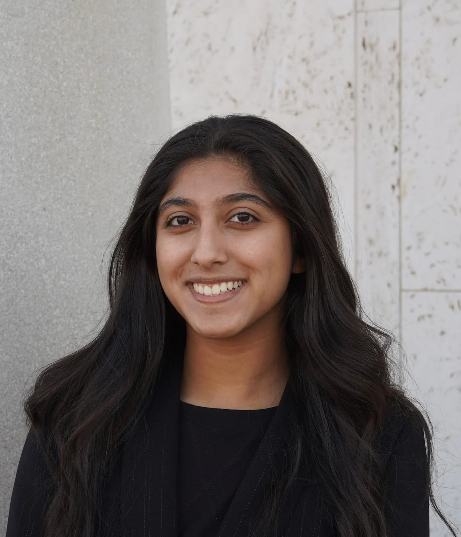 Priyana Manglani headshot: her appearance is that of long dark wavy hair, smiling, against a background of textured gray and white walls.