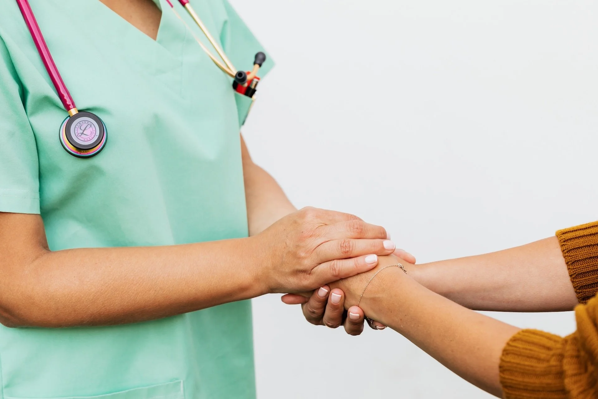 A nurse in green scrubs with a pink stethoscope clasping a patient's hands in a gesture of compassionate care