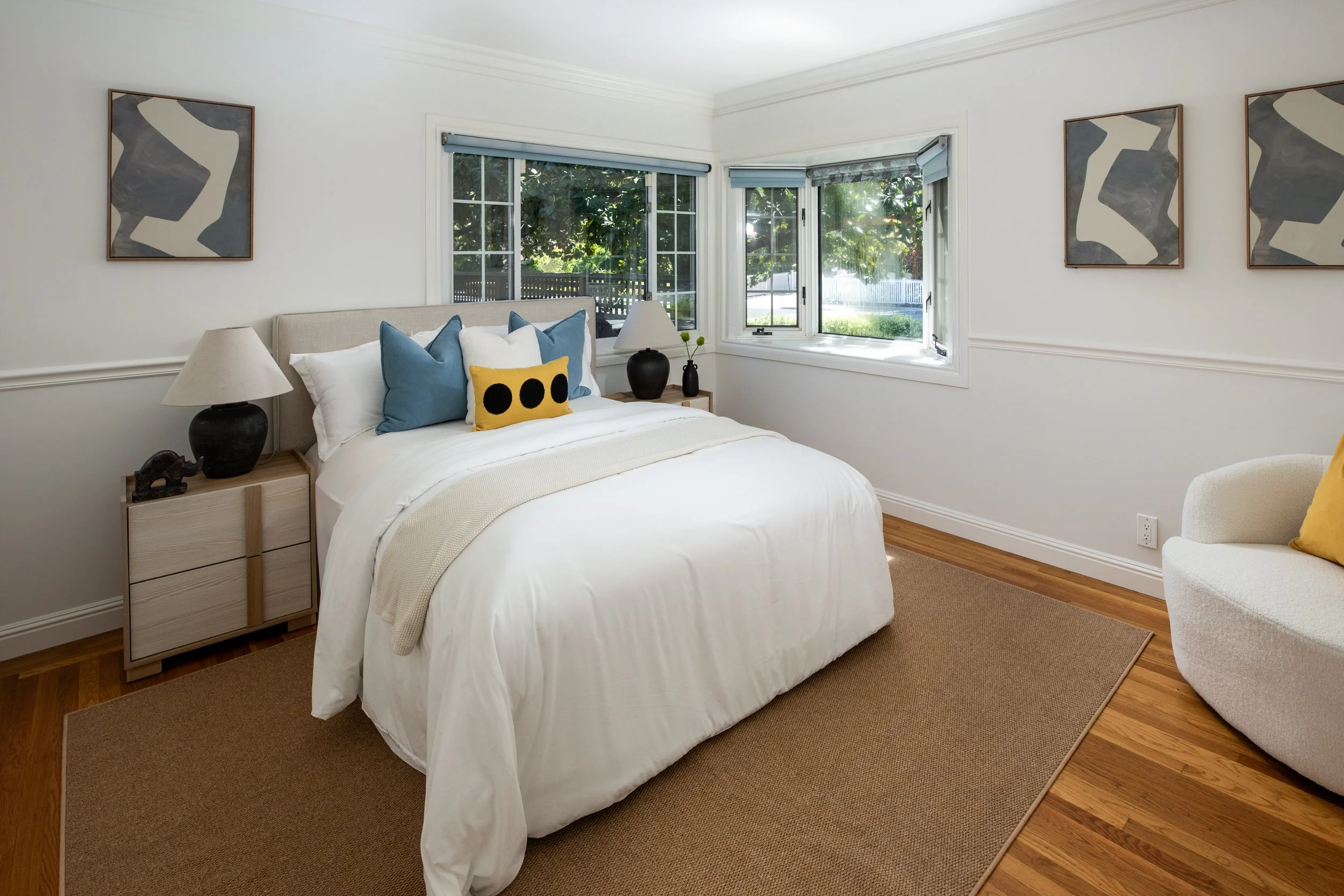 A bedroom with a bed, pillows, nightstand, lamps, artwork, and windows showing greenery outside.