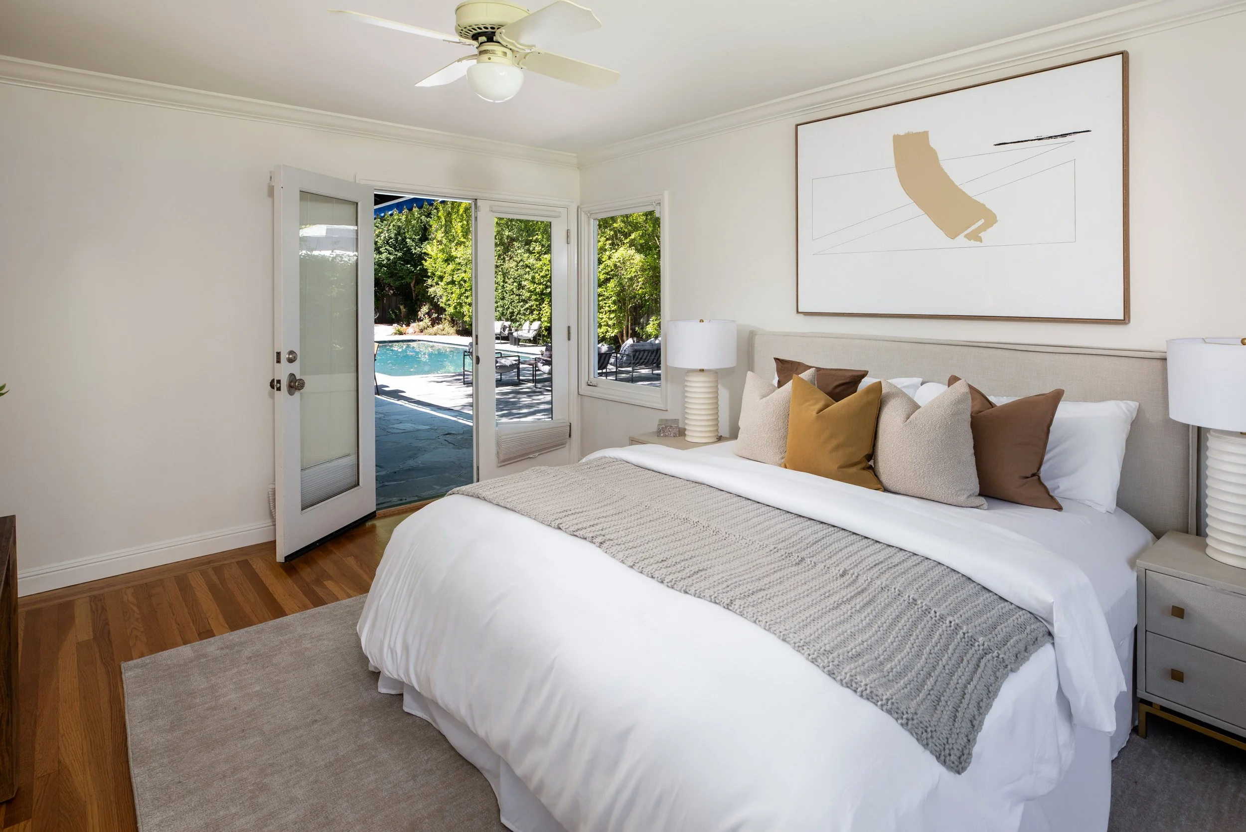A bedroom with a large bed, colorful pillows, and white bedside tables with lamps, open to an outdoor patio with a swimming pool and lush green trees.