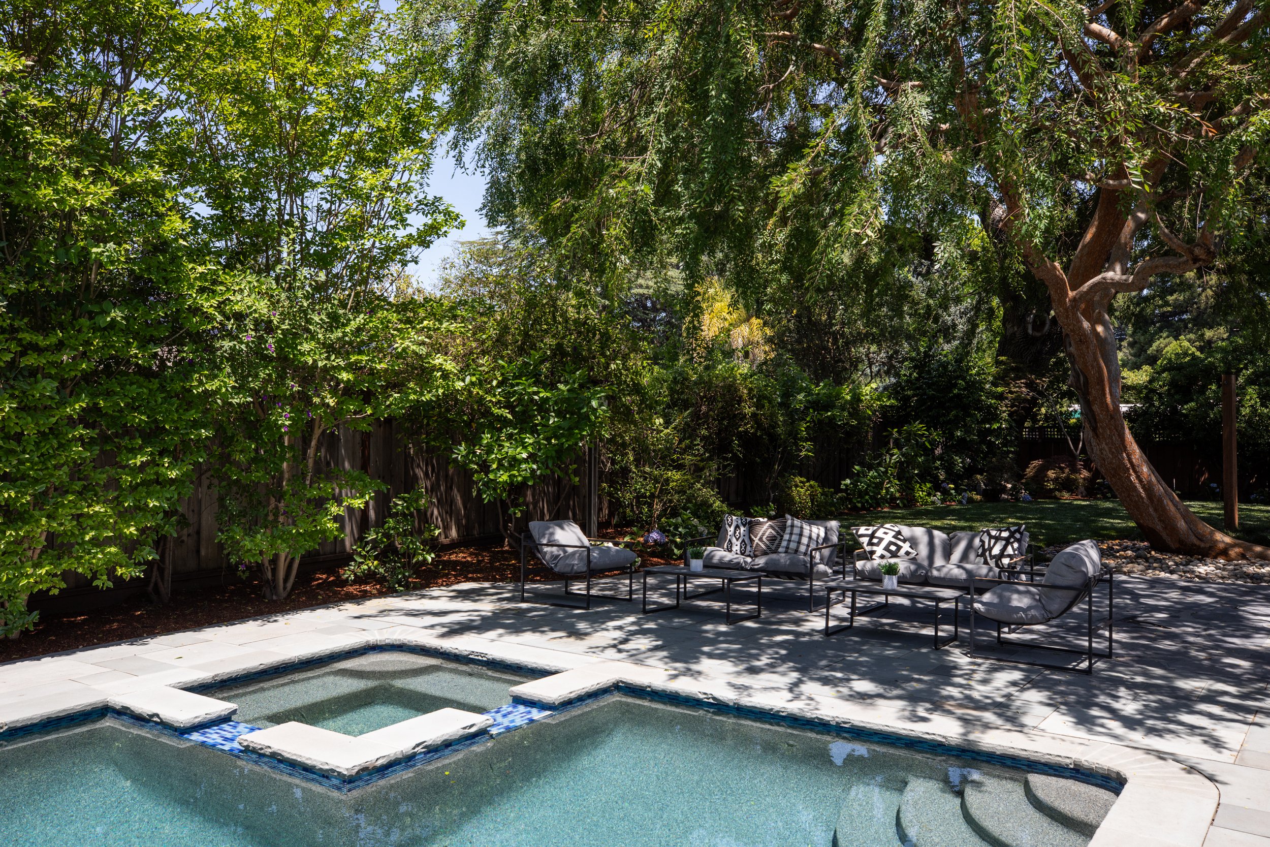 A backyard with a swimming pool, outdoor seating area with lounge chairs and a sofa, surrounded by trees and greenery under a sunny sky.