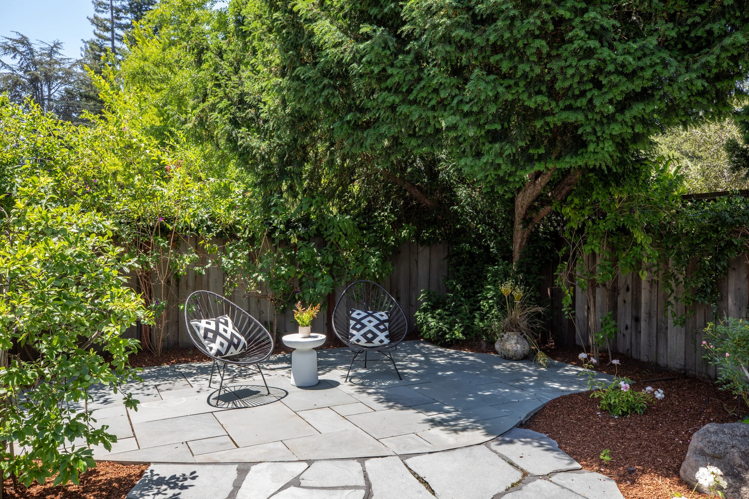 A backyard patio area with two black metal chairs with black and white cushions, a small white table with a potted plant, surrounded by a wooden fence and lush green trees and shrubs.