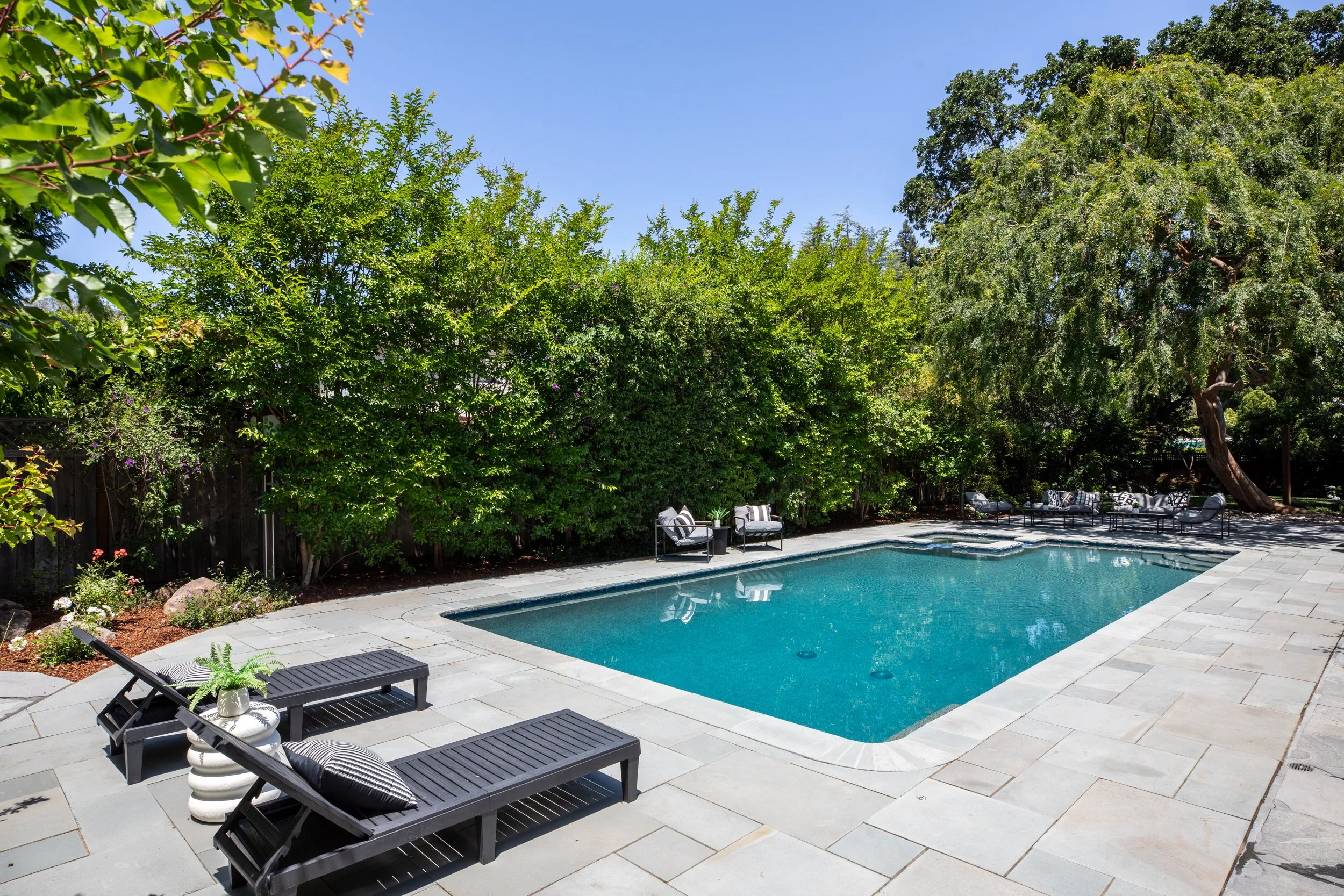A backyard with a swimming pool surrounded by a stone patio, outdoor furniture, and tall green trees and bushes under a clear blue sky.