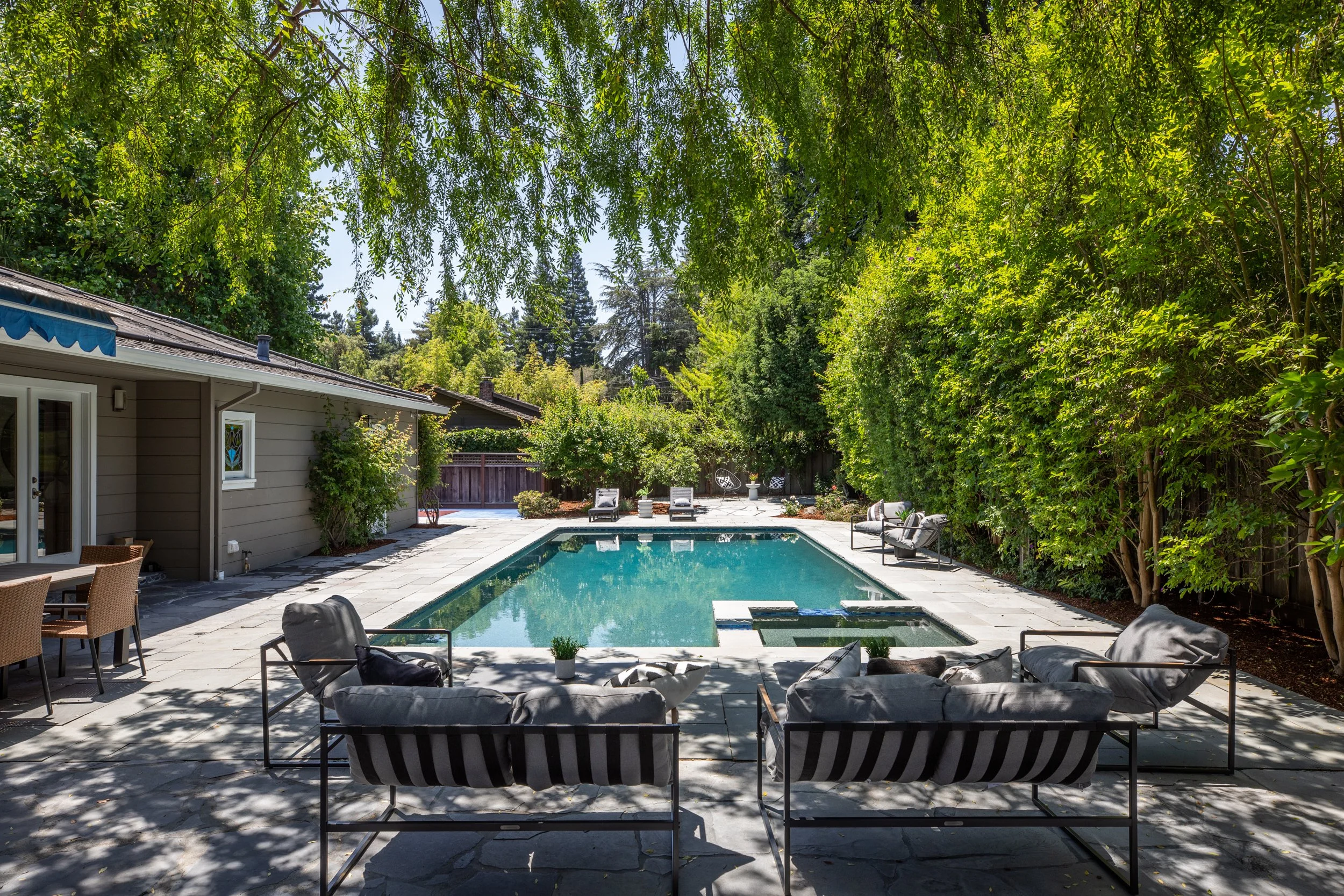 Backyard with a swimming pool surrounded by lounge chairs, lush green trees, and a house with a gray exterior and blue awning.