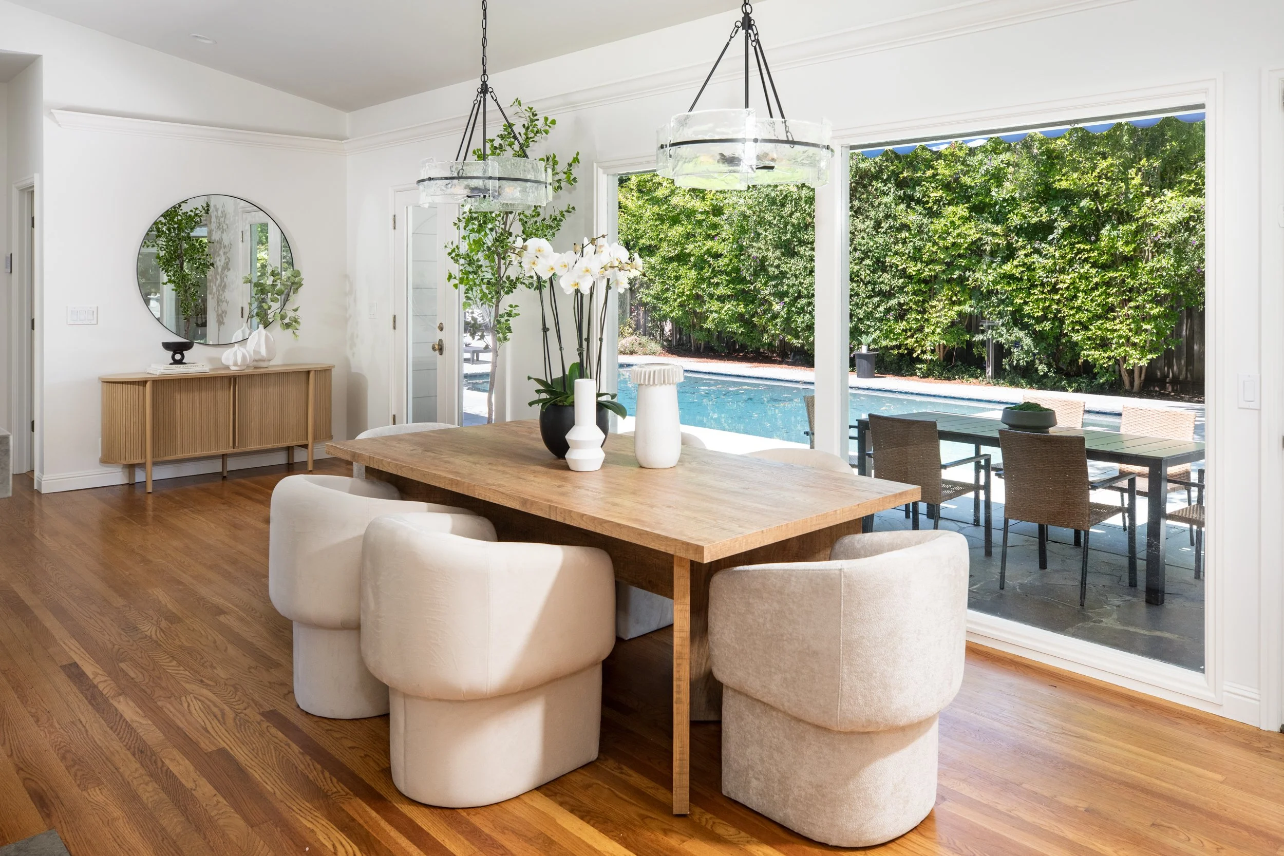 A dining room with a large wooden table surrounded by plush beige chairs, a vase with white orchids, and decorative vases, with large sliding glass doors open to a backyard with a pool and lush green trees.