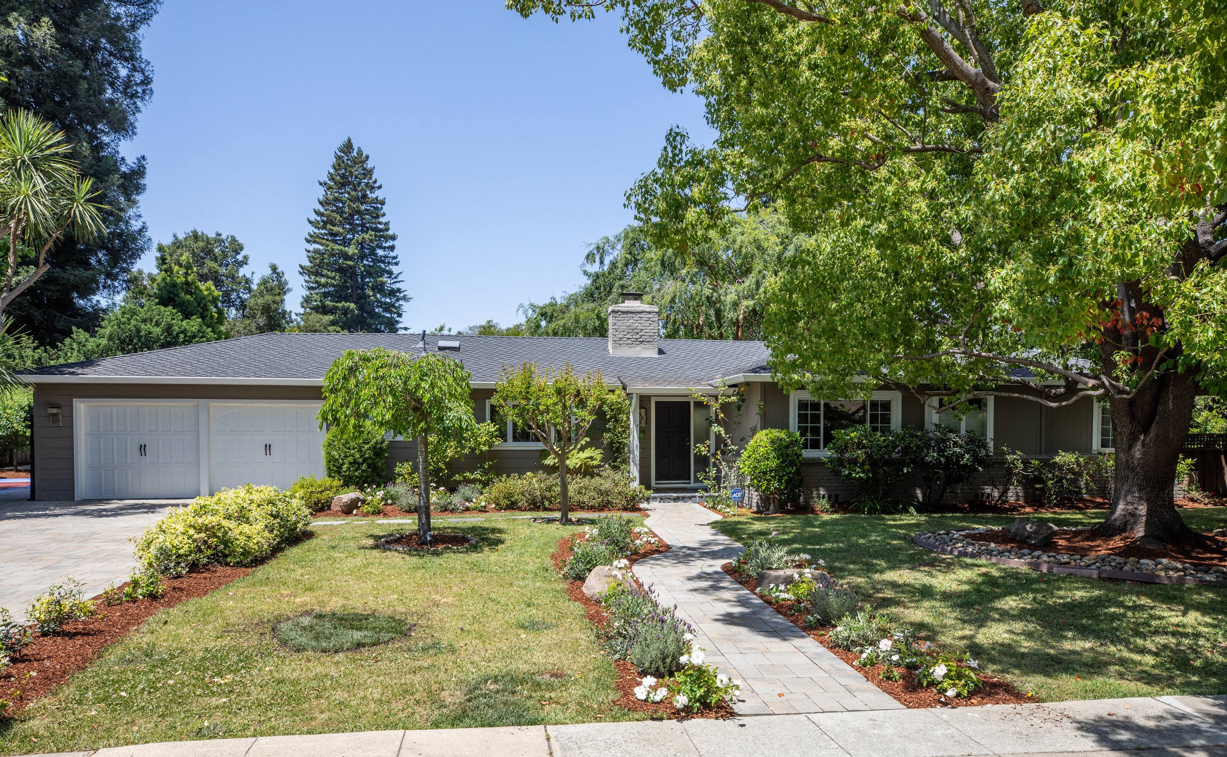 Front view of a house with a gray roof, surrounded by a lush garden and large trees, with a paved walkway leading to the entrance.