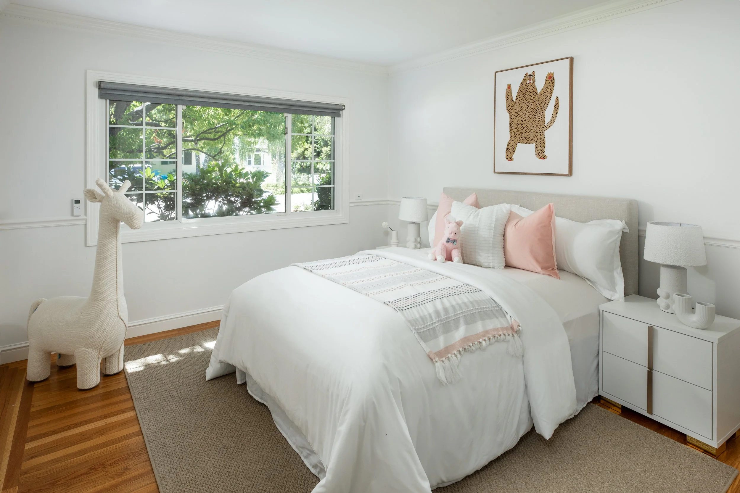 A bedroom with white walls, a large window with gray blinds, a bed with white bedding and pink pillows, and a toy giraffe on the left side. There is artwork of a leopard on the wall above the bed, and two white nightstands with lamps on each side of 