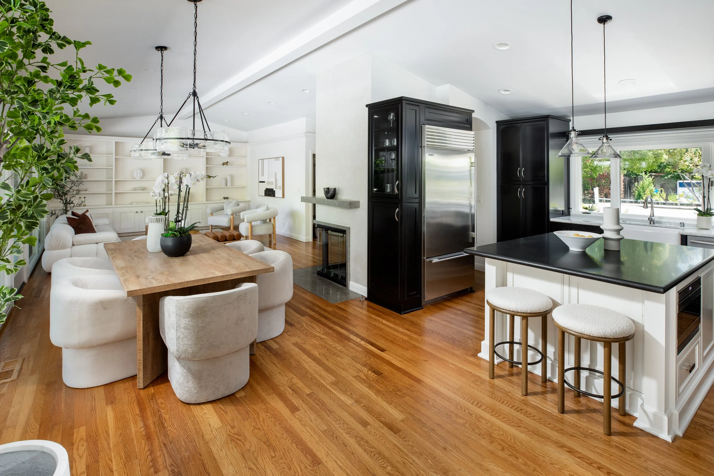 Open-concept kitchen and living room with white walls, hardwood floors, black cabinetry, and large windows showing outdoor greenery. The kitchen features a black countertop with two bar stools, a stainless steel refrigerator, and pendant lighting. Th