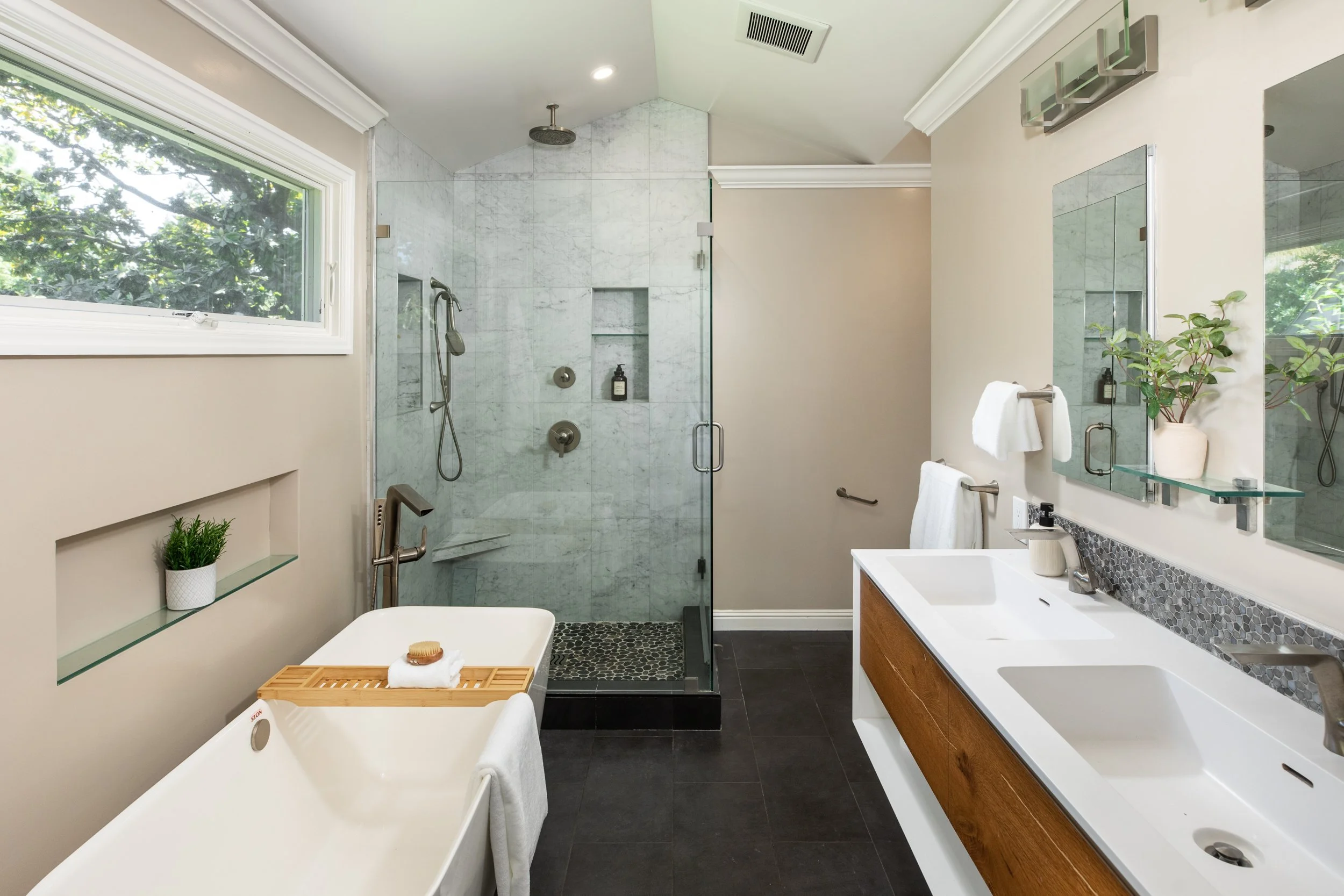 Modern bathroom with beige walls, a large window, a glass-enclosed walk-in shower with marble tiles, a white bathtub with a wooden tray, a vanity with a sink, mirror, and cabinets, potted plant, soap, and towels.