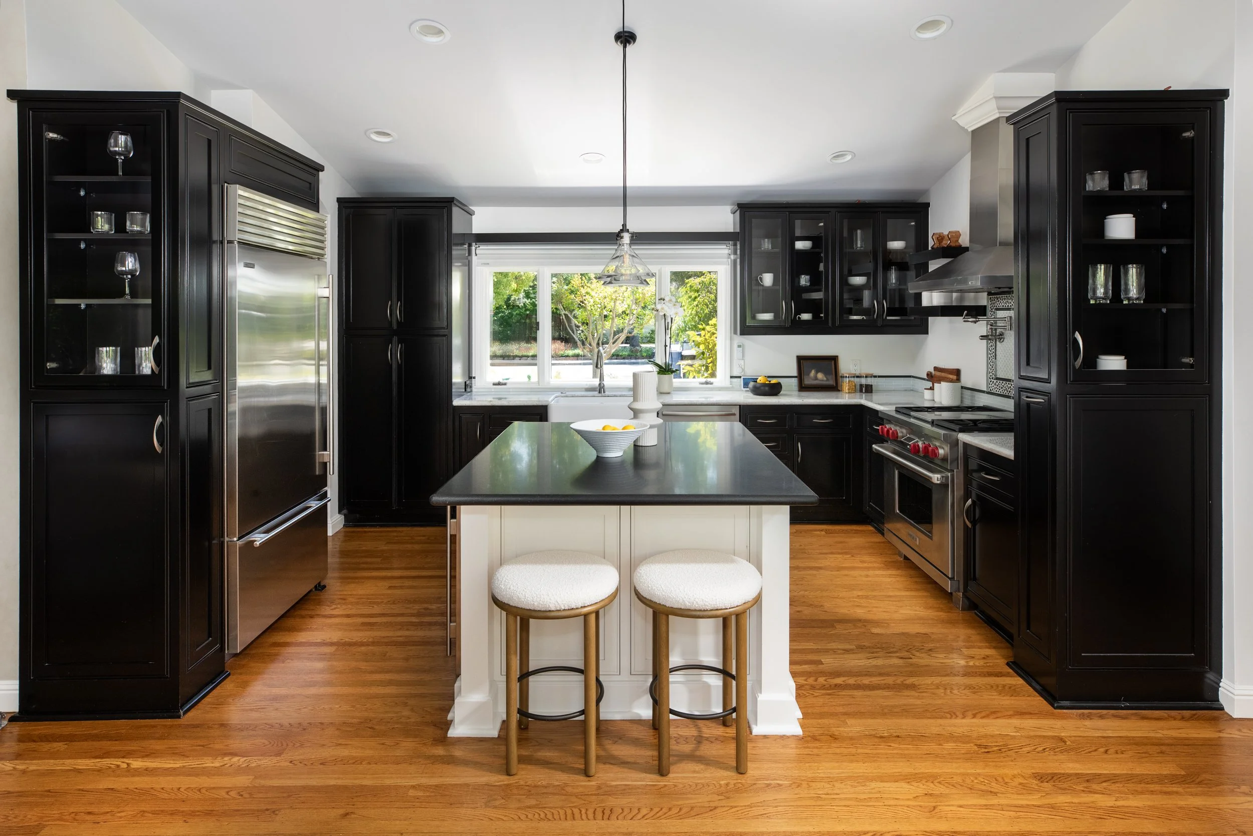A modern kitchen with black cabinets, a white island with two stools, hardwood floors, and a large window overlooking greenery.