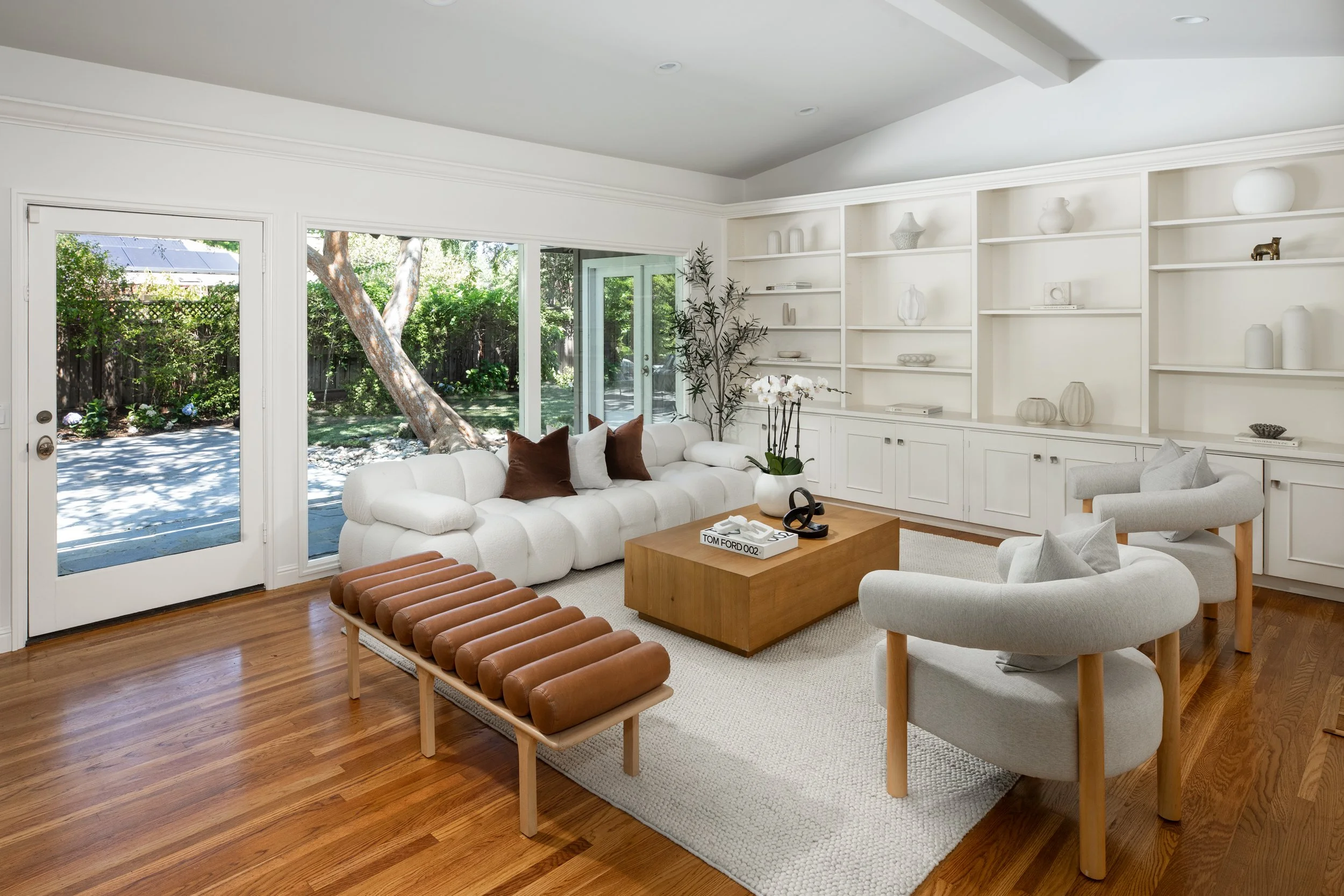 Bright living room with white walls, wooden floors, large sliding glass doors leading to a garden, and a built-in white bookshelf. Furniture includes a white tufted sofa, light gray armchairs, a wooden coffee table, and a brown leather bench. Decor f