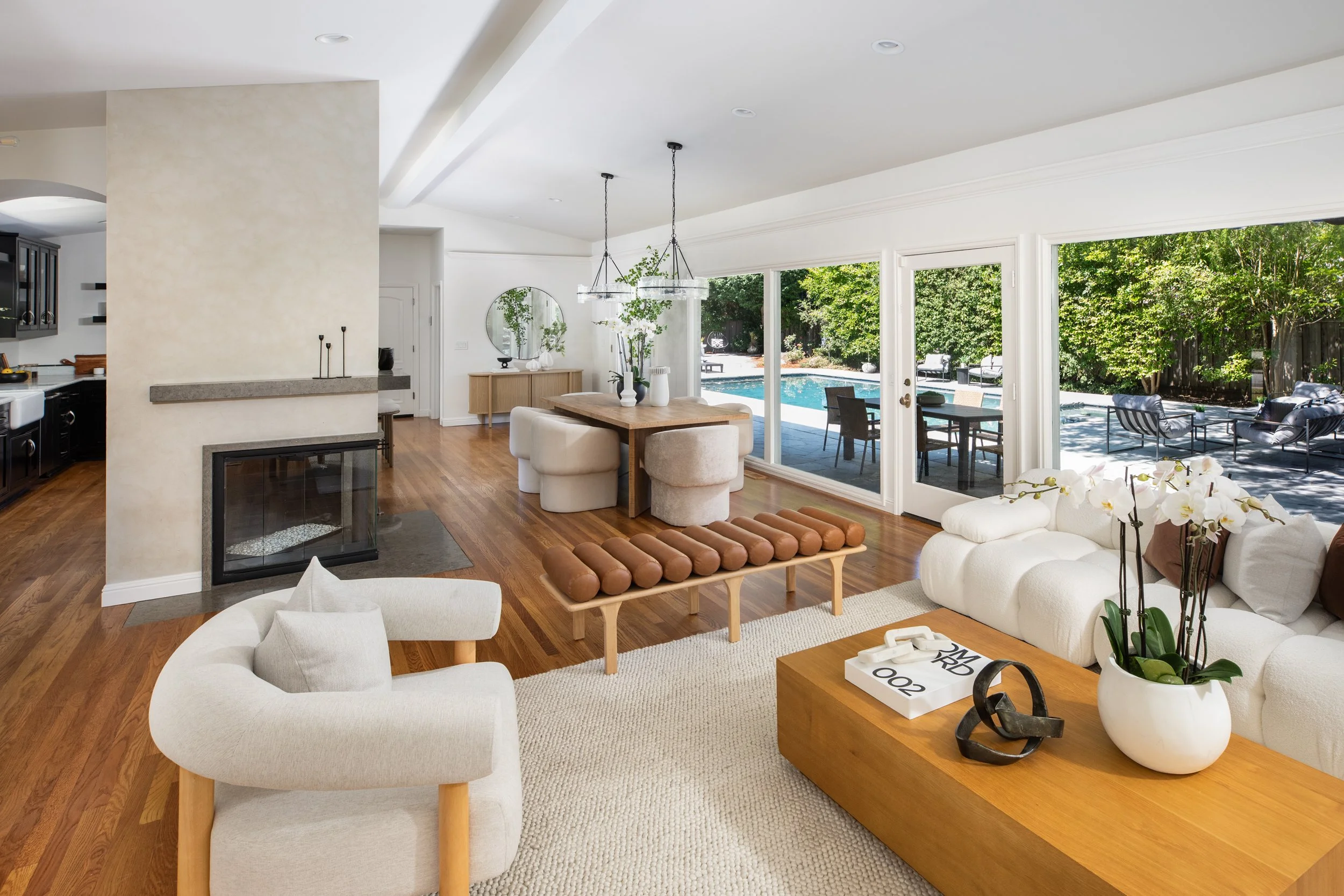 Bright modern living room with white sofas, round armchair, wooden coffee table with decorative objects, and large sliding glass doors leading to outdoor patio and swimming pool surrounded by green trees.