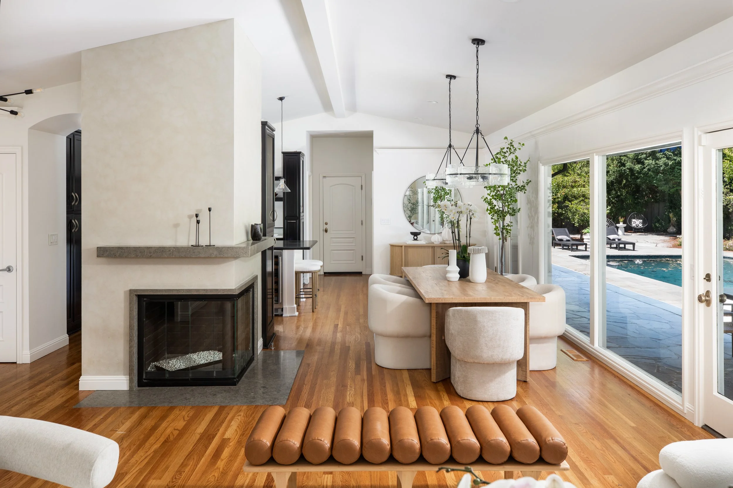 Open-concept living and dining area with hardwood floors, large glass doors leading outside to pool and patio, white walls, modern black pendant lights, and a mix of contemporary and rustic furniture.