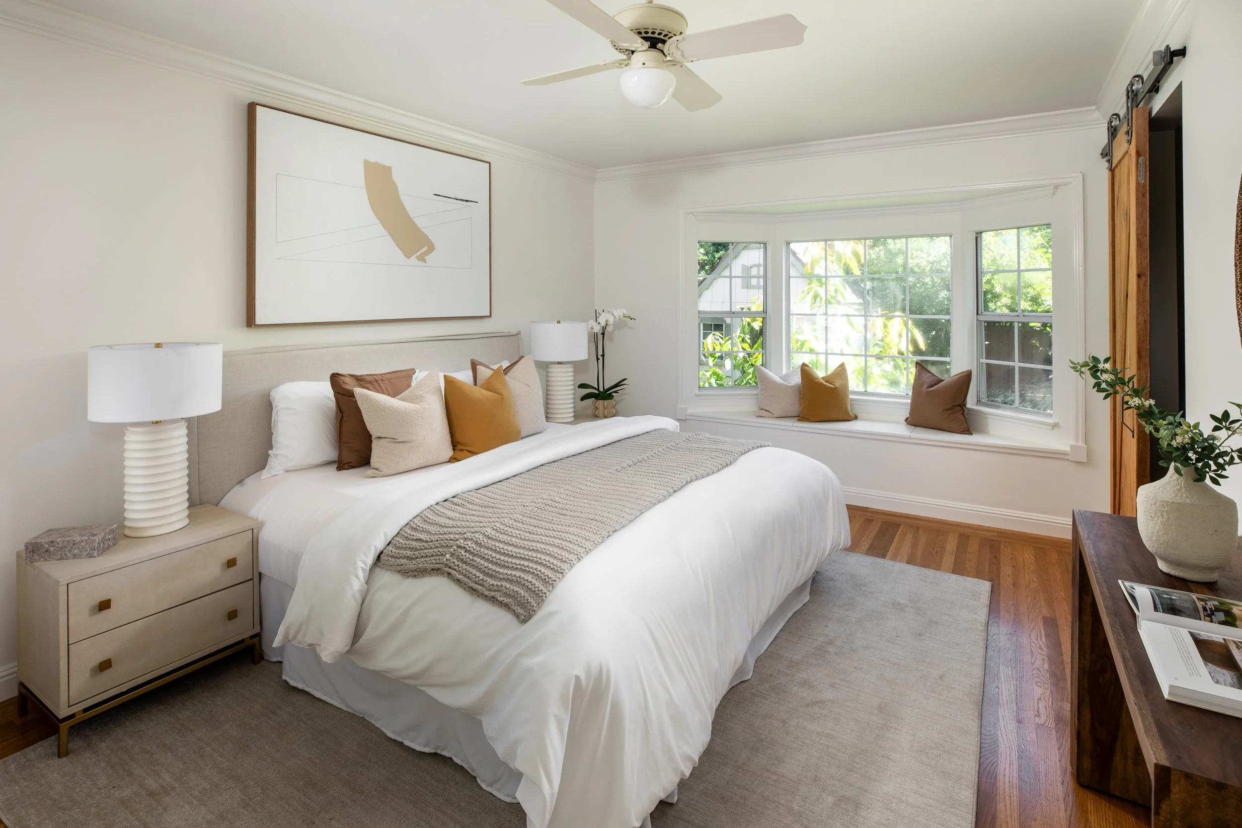 A cozy bedroom with a large bed decorated with multiple pillows in neutral tones, two white bedside lamps, and a window seat with cushions overlooking greenery outside. A ceiling fan hangs from the ceiling, and a minimalist wall art piece adorns the 