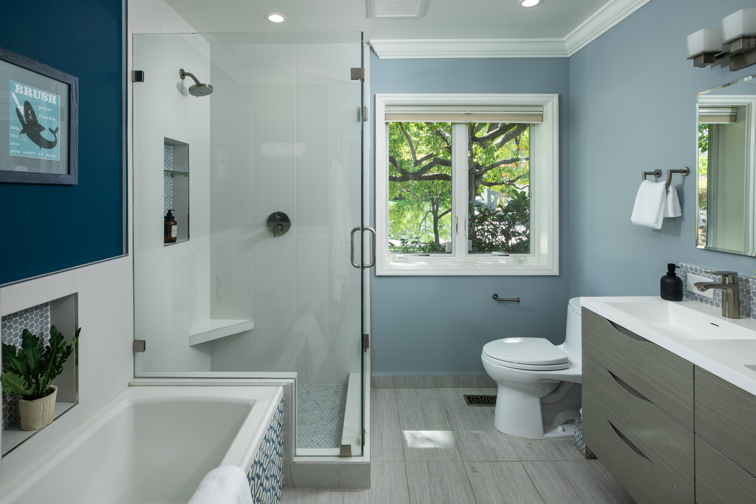 Bathroom with a glass-enclosed shower, a white toilet, a vanity with a sink, a mirror, and a window showing green trees outside.