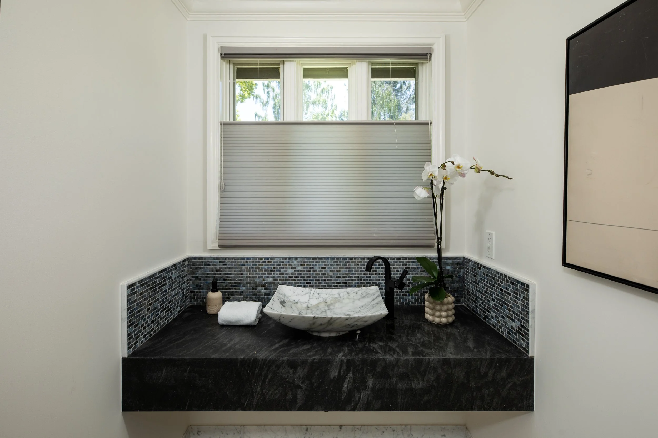 Bathroom with a window, a white orchid, a marble sink, black faucet, soap dispenser, a rolled towel, dark tiled backsplash, black countertop, white walls, and framed mirror.