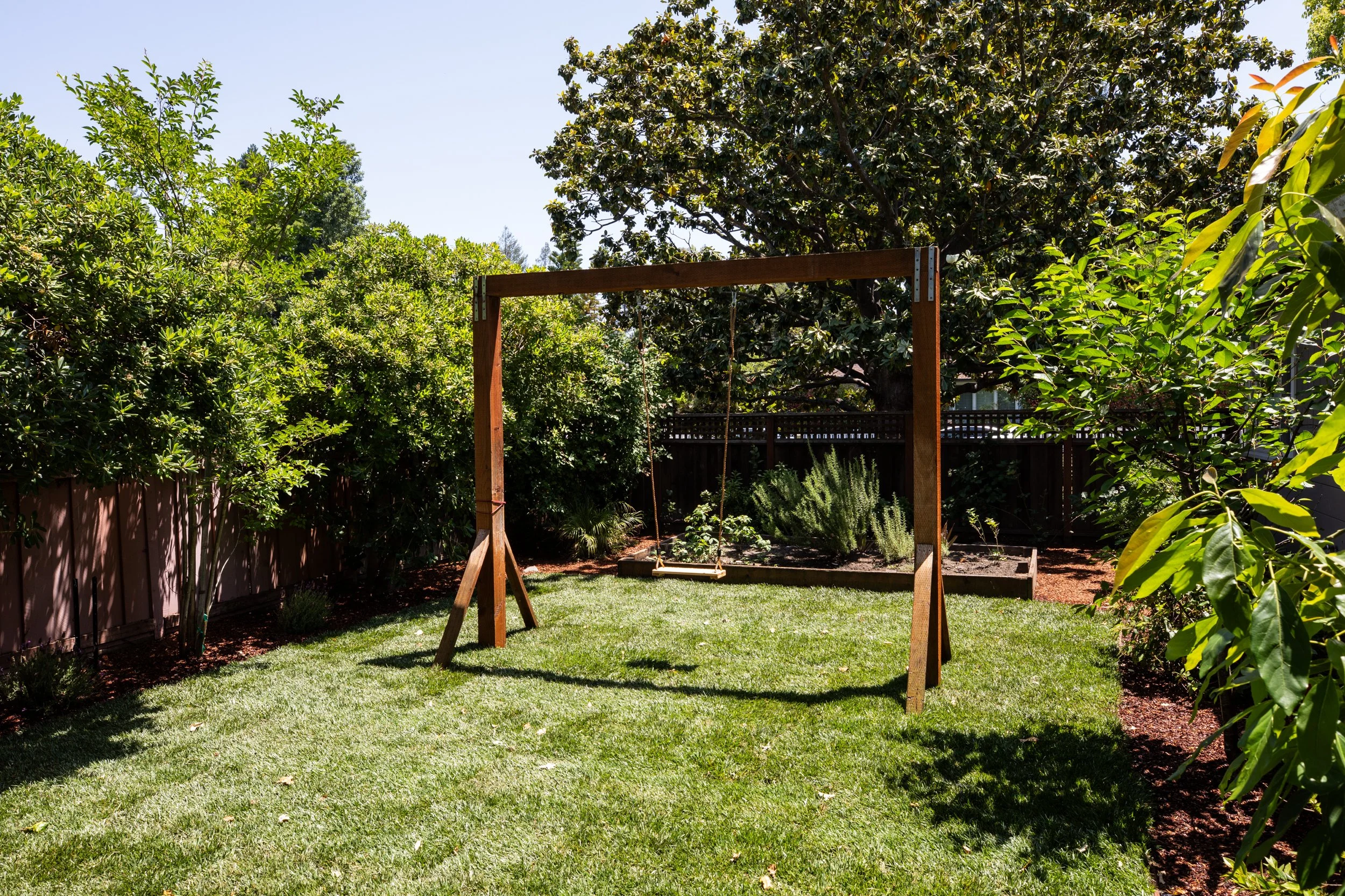 A backyard with a swing set frame, surrounded by lush green trees and shrubs, with a well-maintained lawn and a clear blue sky.