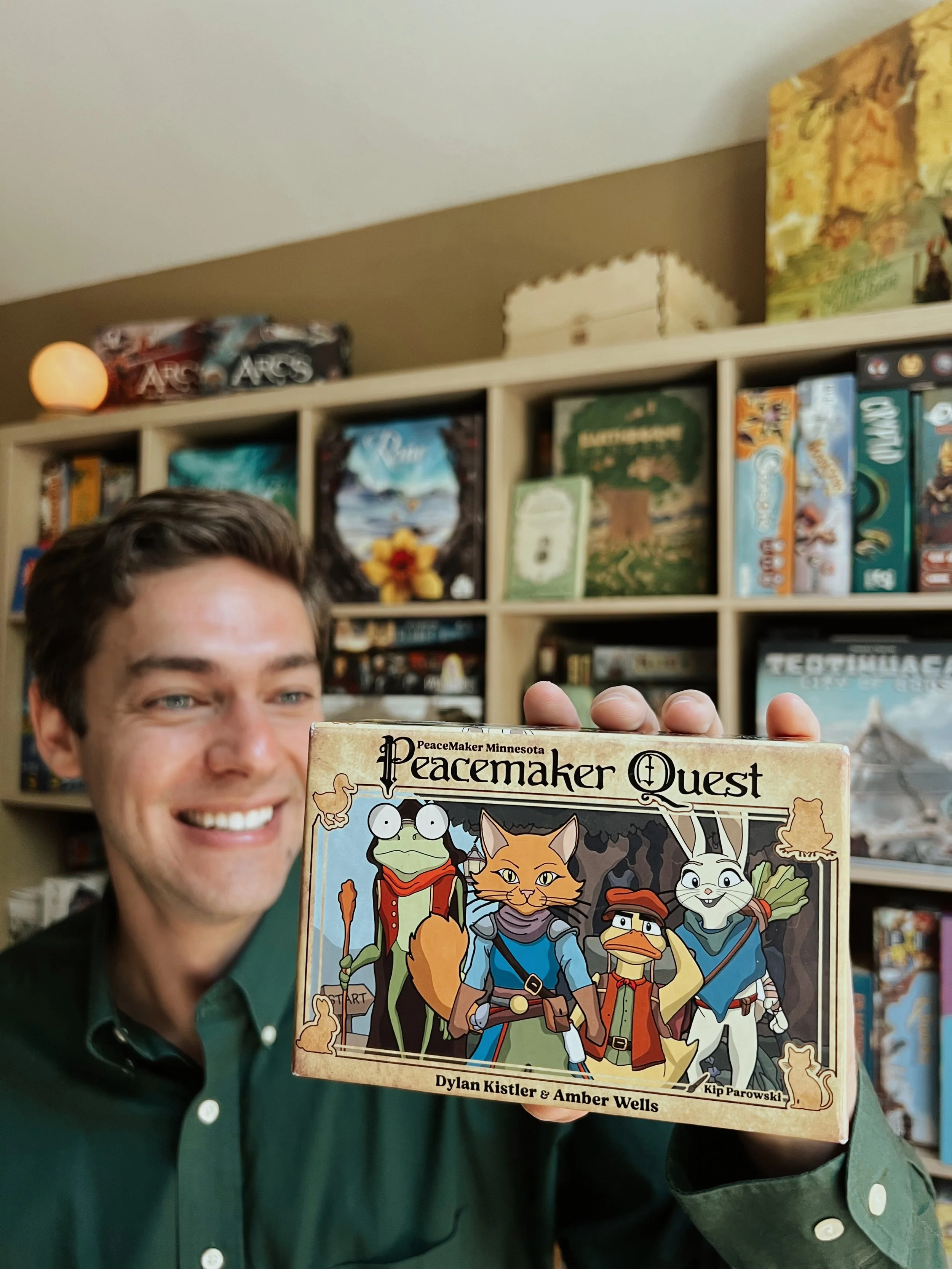 Dylan Kistler, Founder of Kistler Games, smiling and holding a board game he developed called 'Peacemaker Quest'. The background shows shelves filled with various board games.