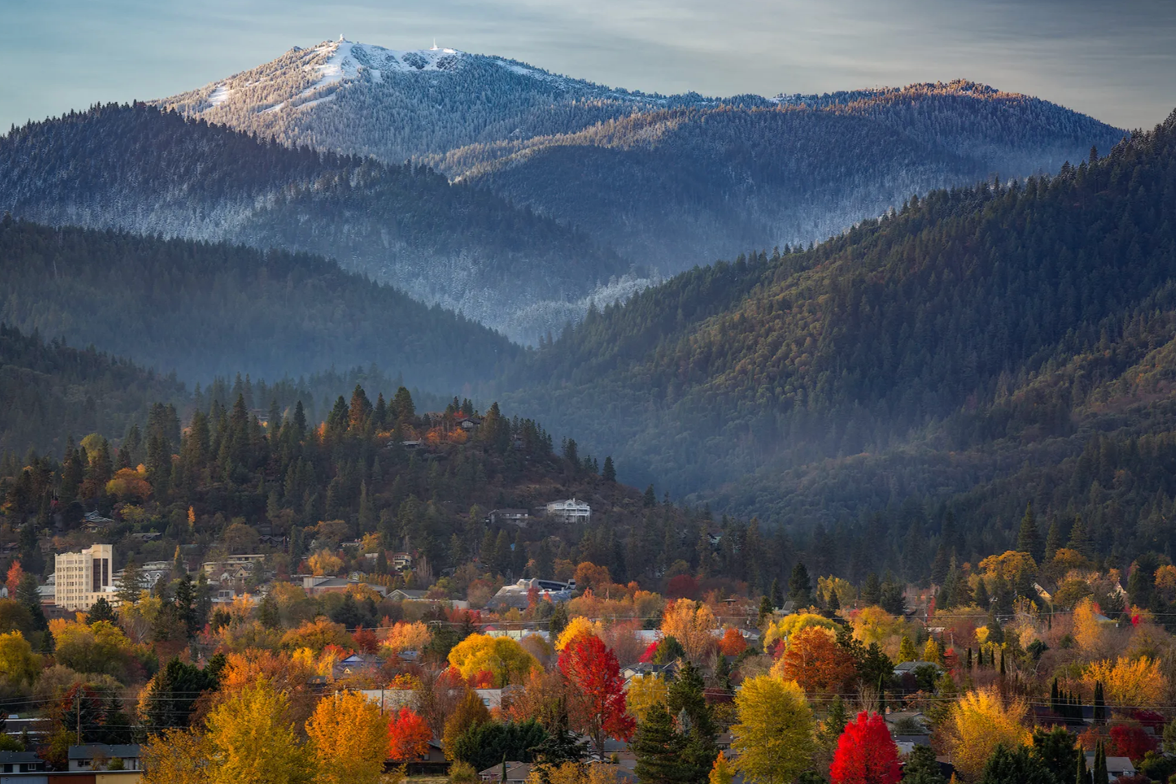 A cityscape with mountains in the background during fall, showing colorful trees in red, yellow, and orange, and houses and buildings in the city below.