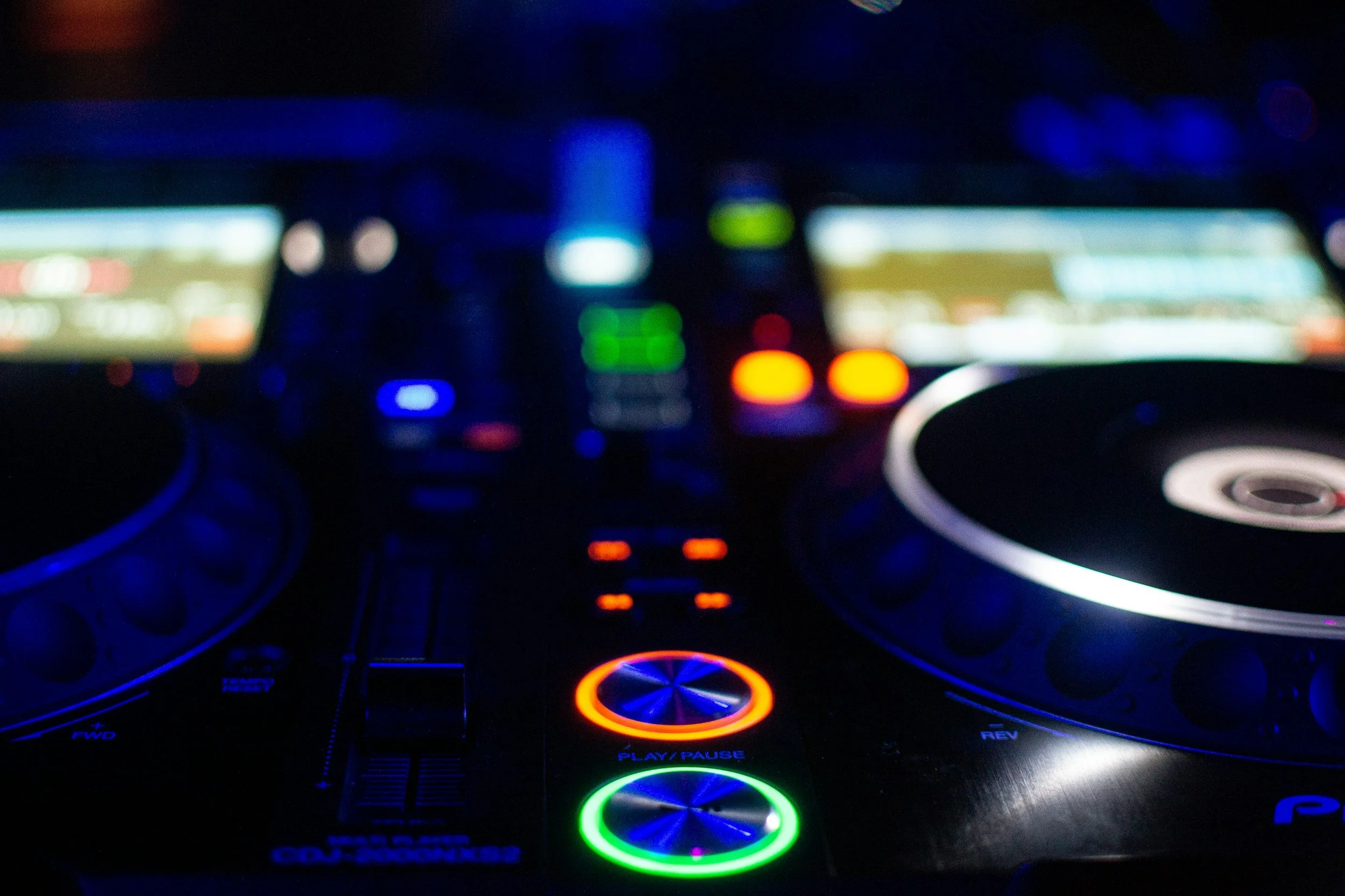 A close-up of a DJ mixer and turntables with colorful illuminated controls and screens in a dark environment.