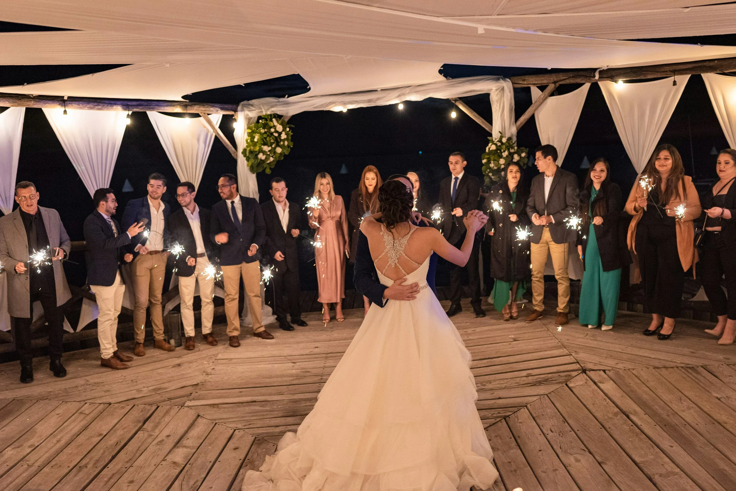 A bride and groom dancing at their wedding reception, with guests standing in a semi-circle around them holding sparklers, under a decorated canopy at nighttime.