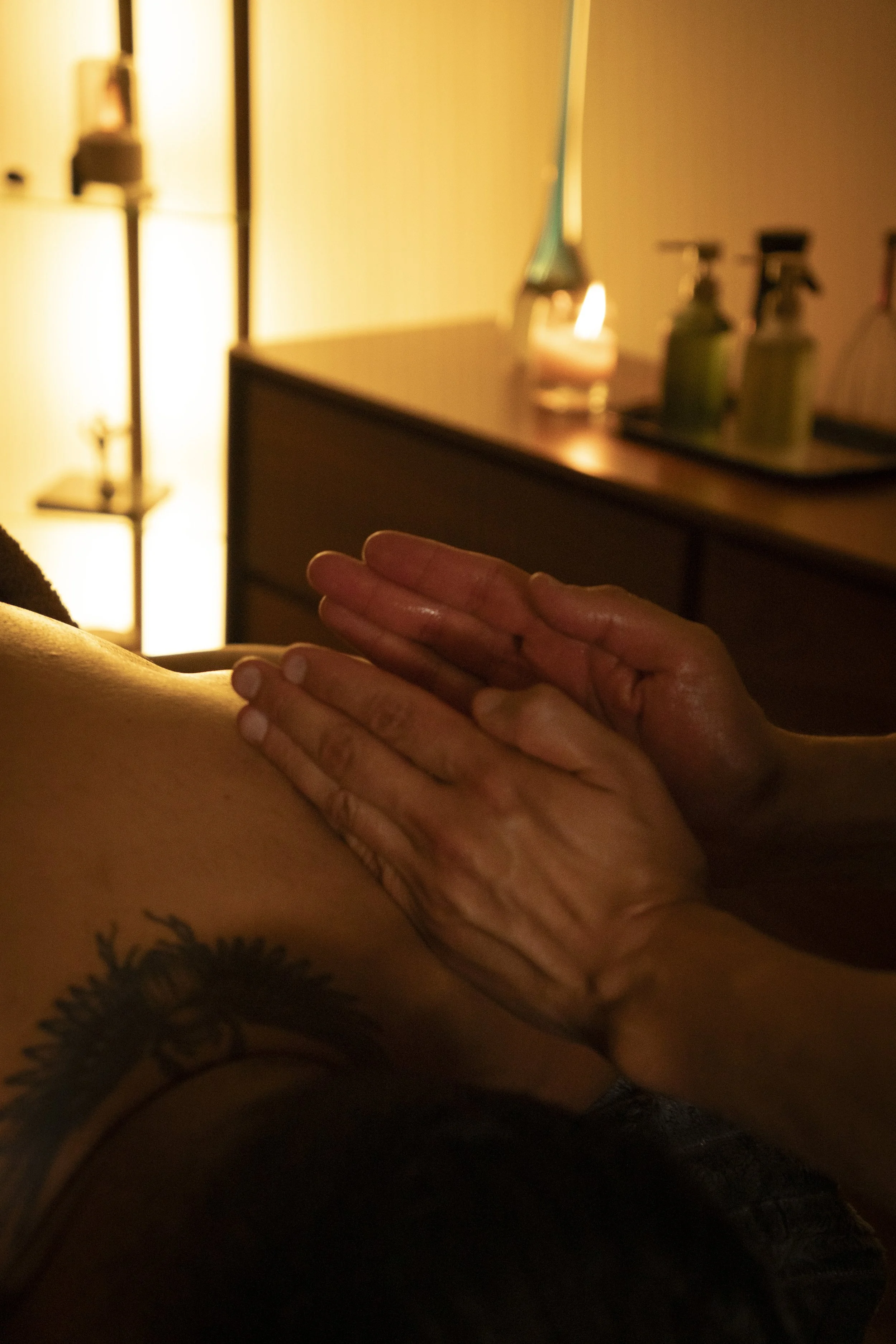 Close-up of a person receiving a massage, focusing on their back, with hands applying pressure, in a warm, softly lit room with candles and bottles of massage oil in the background.