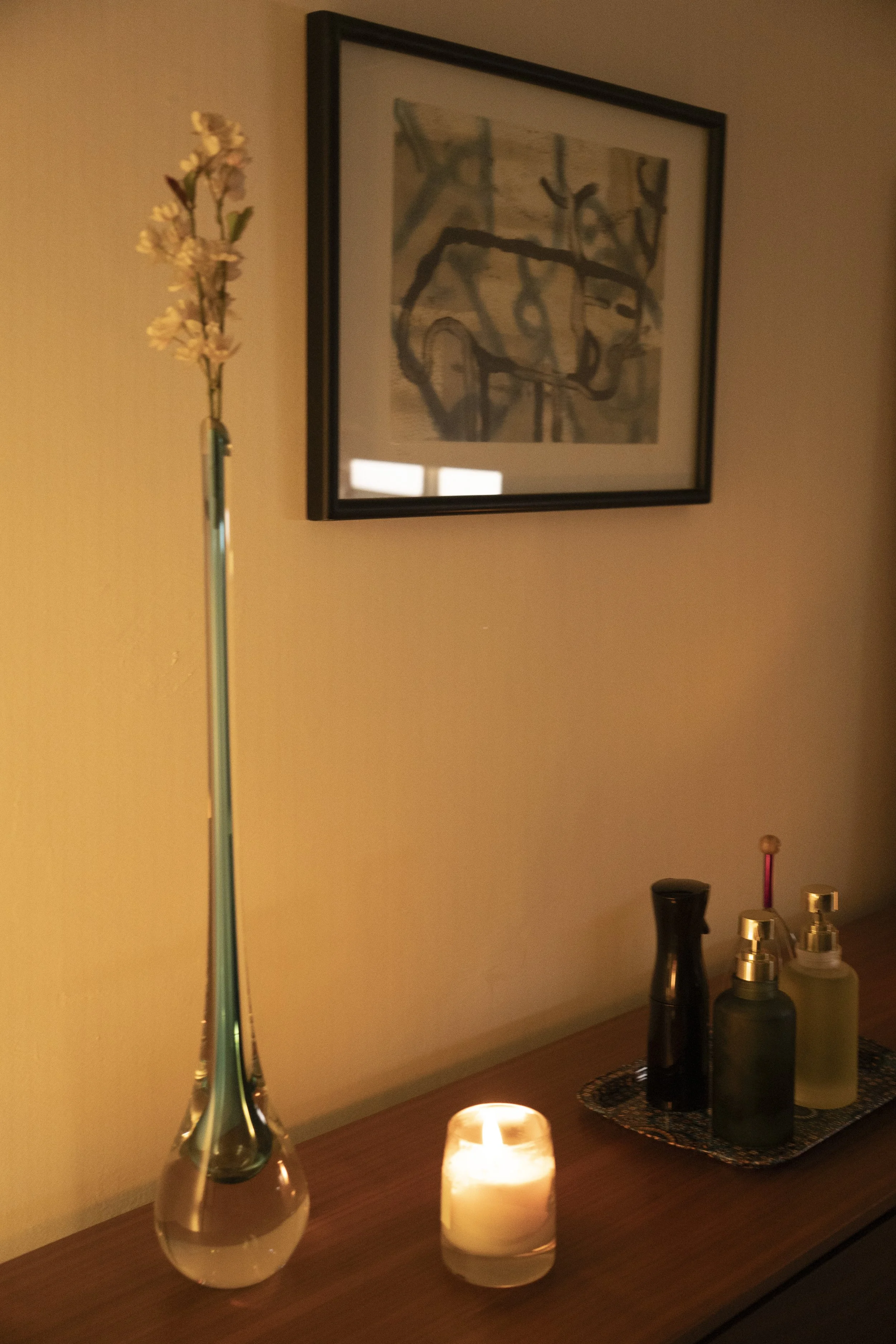 A wooden surface with a glass vase holding a single pink and white flower, a lit candle, and a black tray with various bottles and containers. A framed abstract artwork hangs on a beige wall behind the surface.