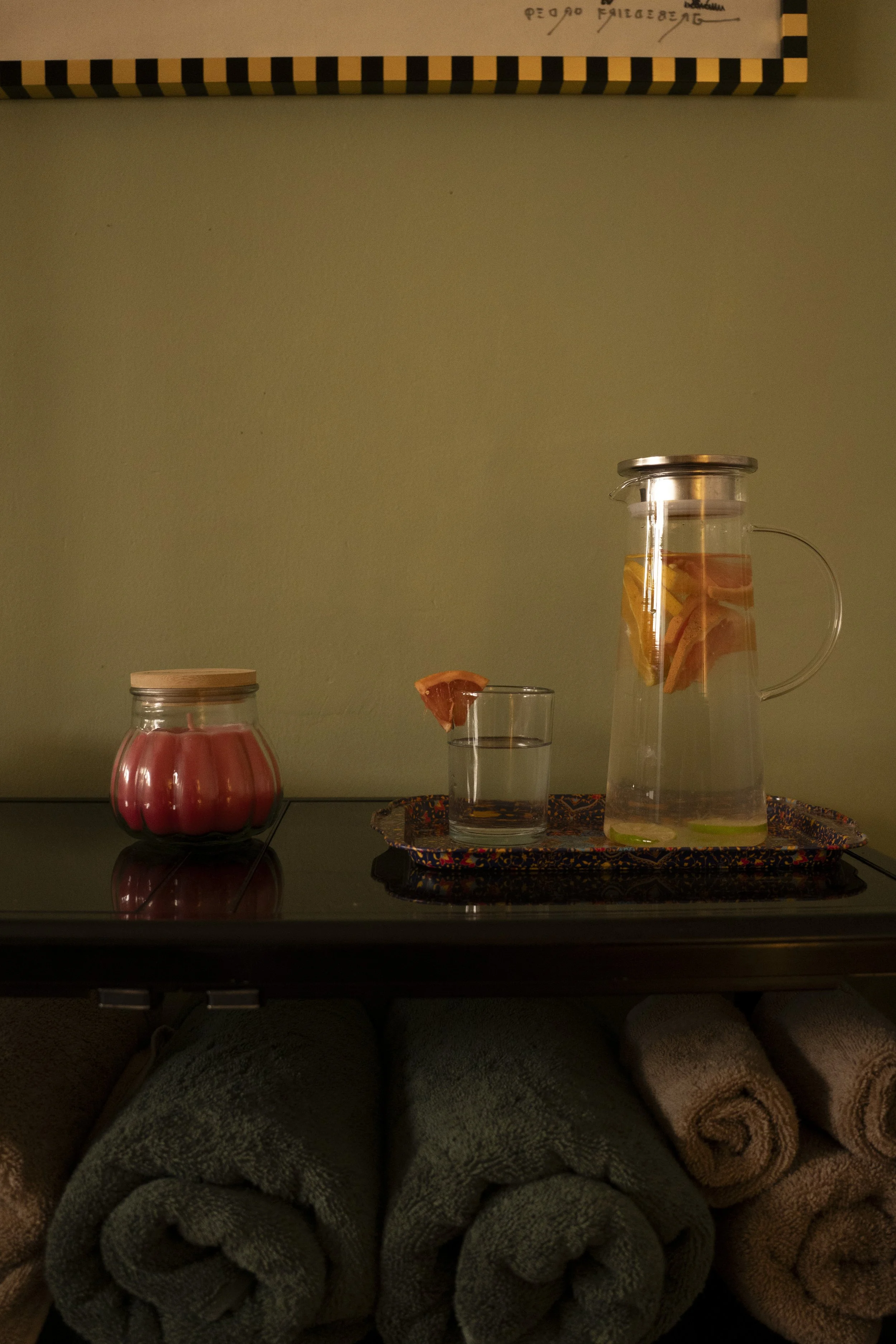 A scene with a glass pitcher filled with water and slices of citrus fruit, a glass with a slice of grapefruit, a jar with a lid, and a tray of rolled towels on a dark surface against a plain wall.