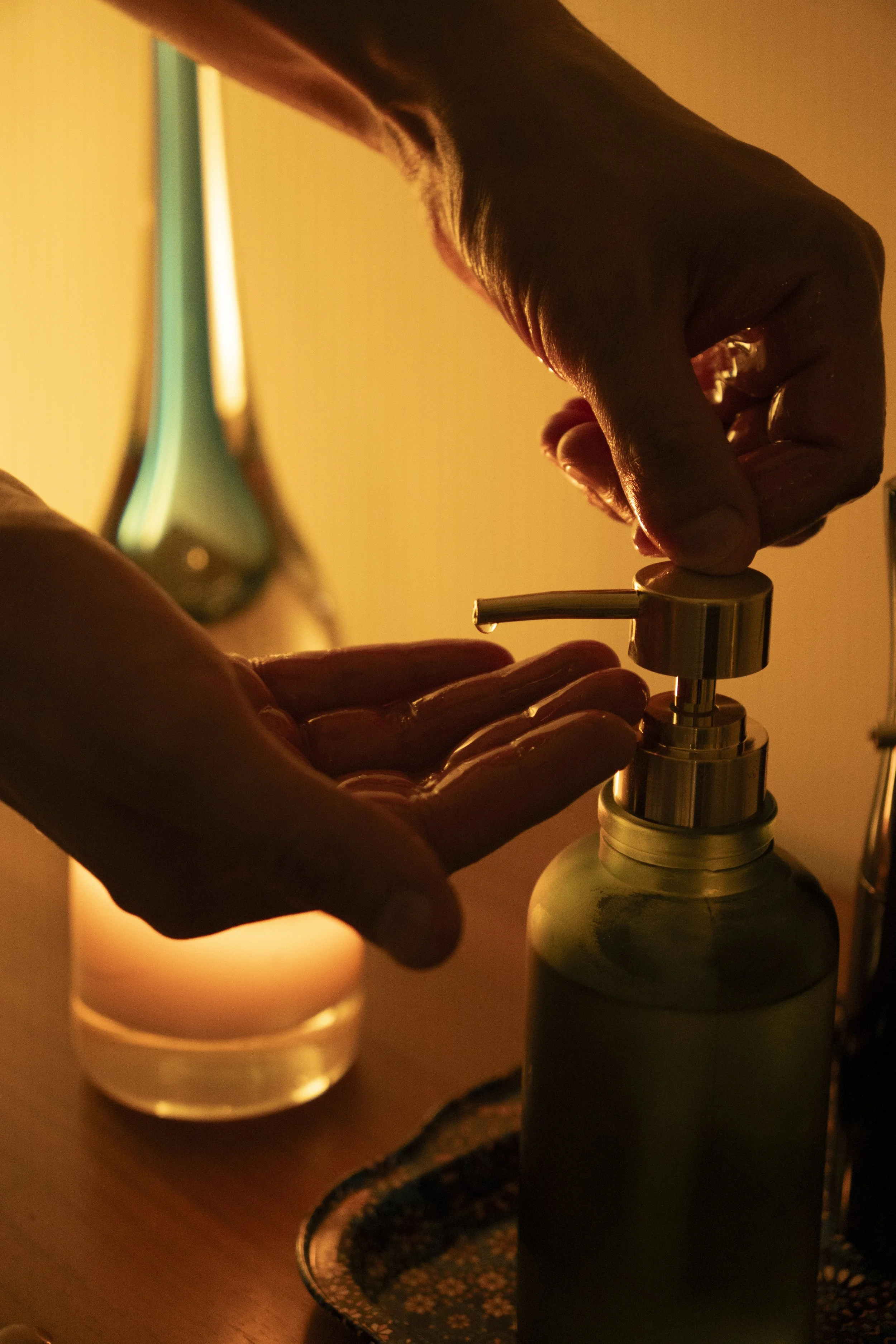 A person pressing a pump dispenser on a bottle, with a glass of water and a small container nearby, in warm lighting.