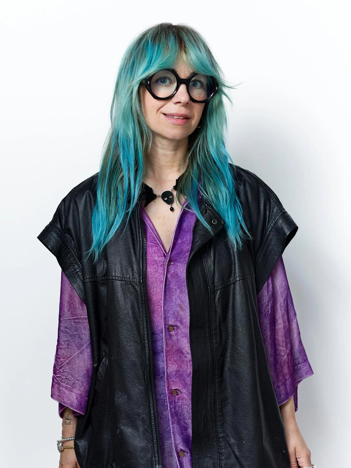 A colorful clay art figurine of a person with teal and blue hair, wearing large round glasses, a skull necklace, a purple tie-dye shirt, and black vest and pants, standing against a white background.