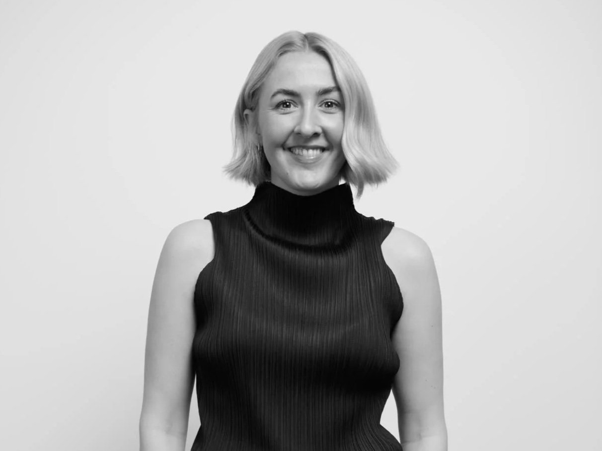 a woman with blonde hair, wearing a black jacket with white buttons and matching black pants, standing against a white background.
