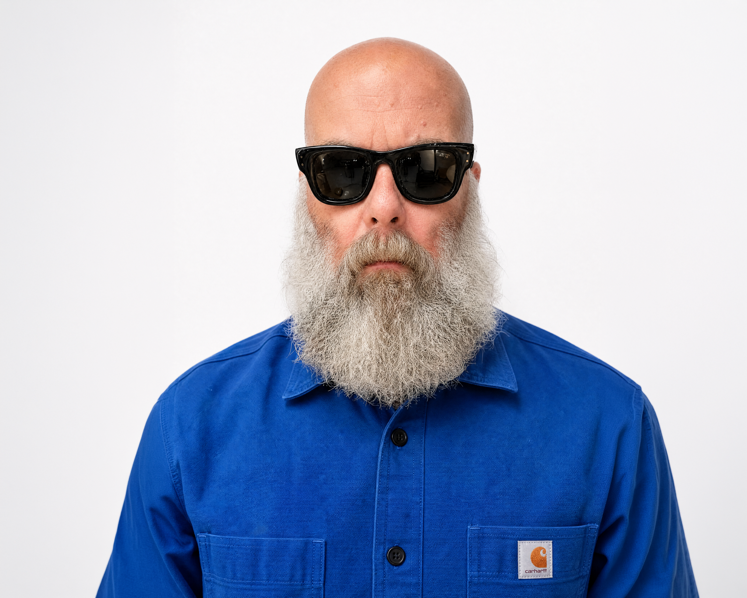 a man with a bald head, white beard, wearing sunglasses, a dark t-shirt, blue jeans, and red, white, and black sneakers, standing against a plain white background.