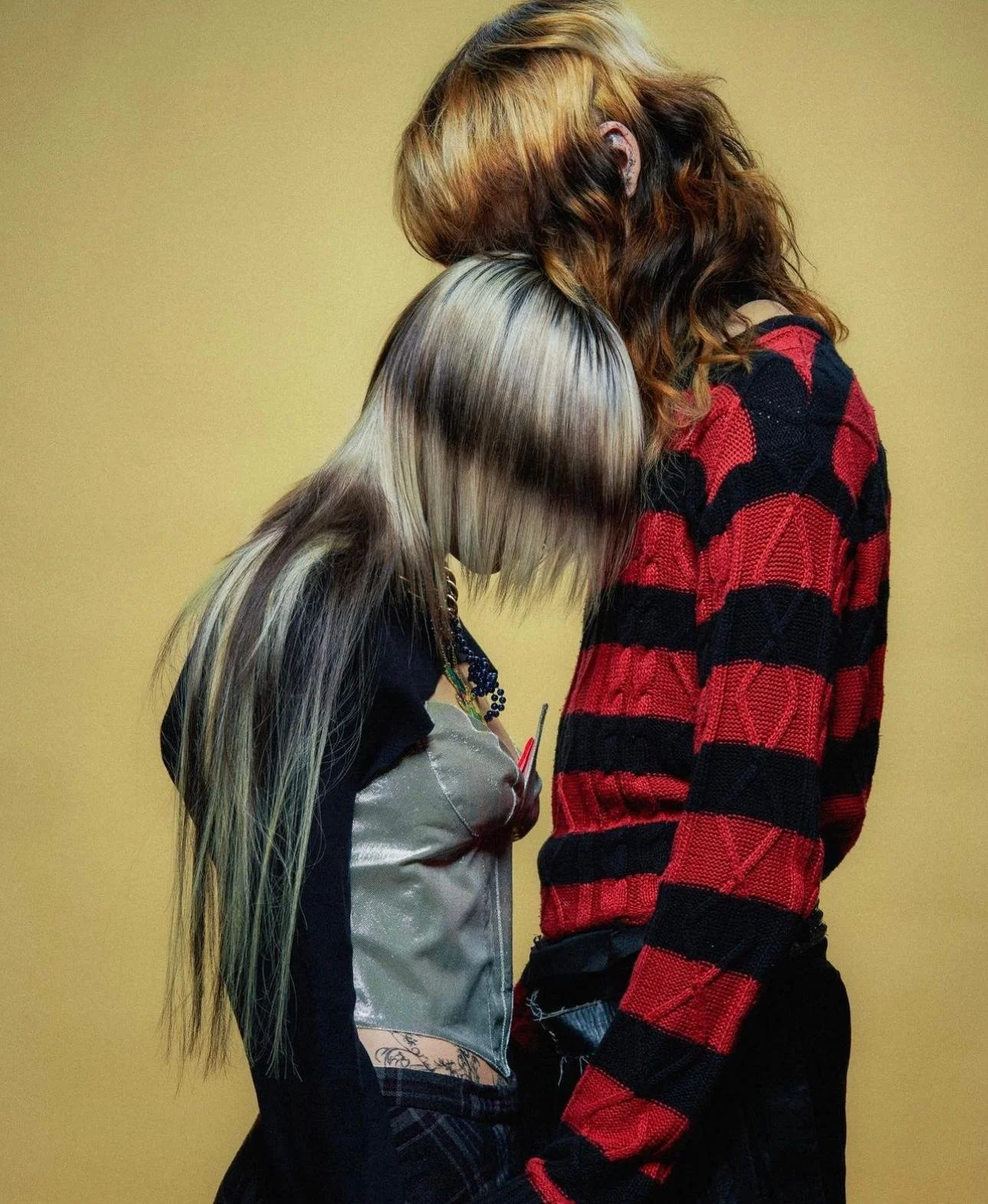 Two women standing closely with their heads touching, one with light blonde hair and the other with red wavy hair, against a plain yellow background.