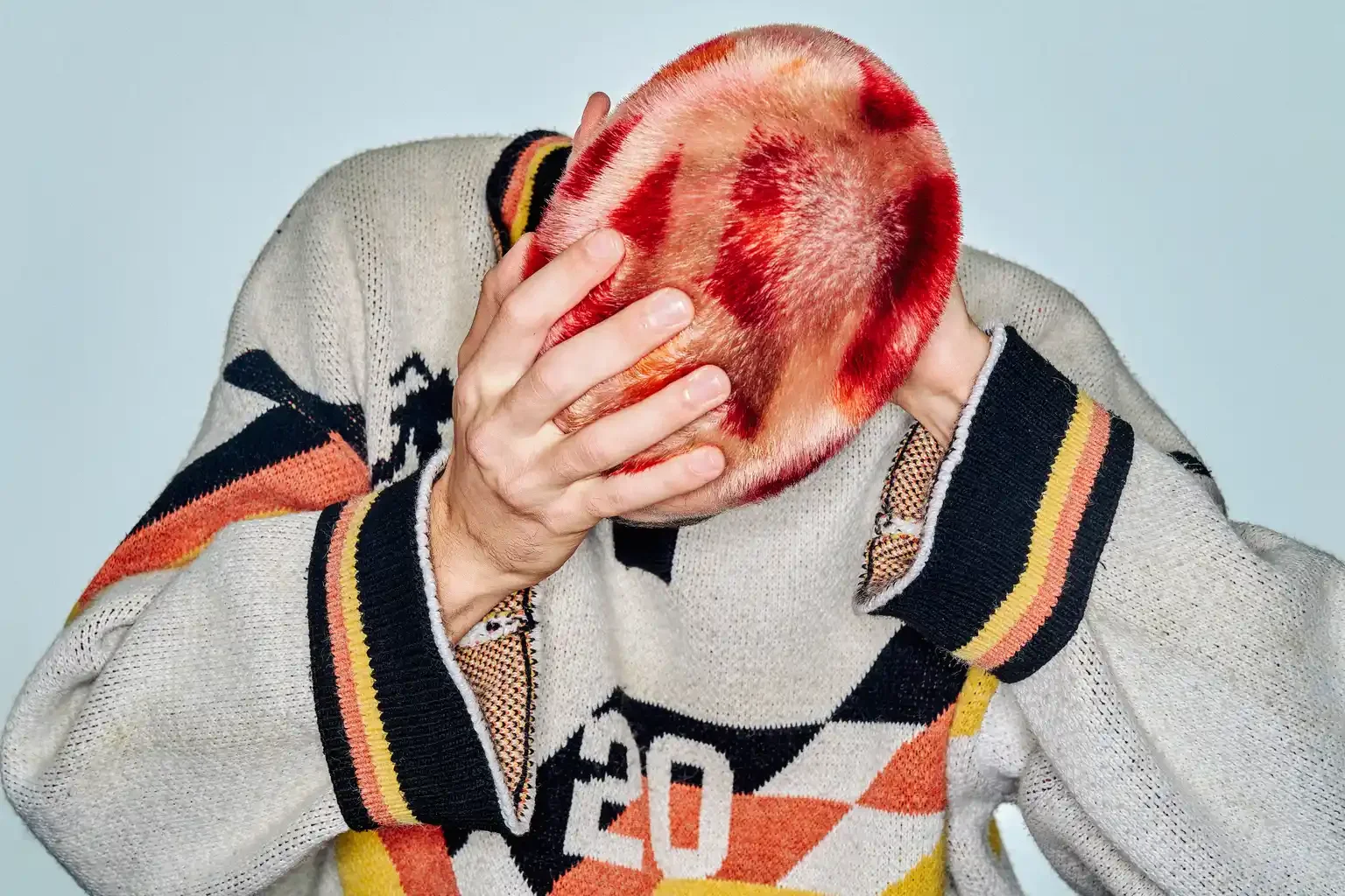 Person with dyed red hair holding their head with both hands, wearing a gray sweater with black, orange, and yellow patterns.