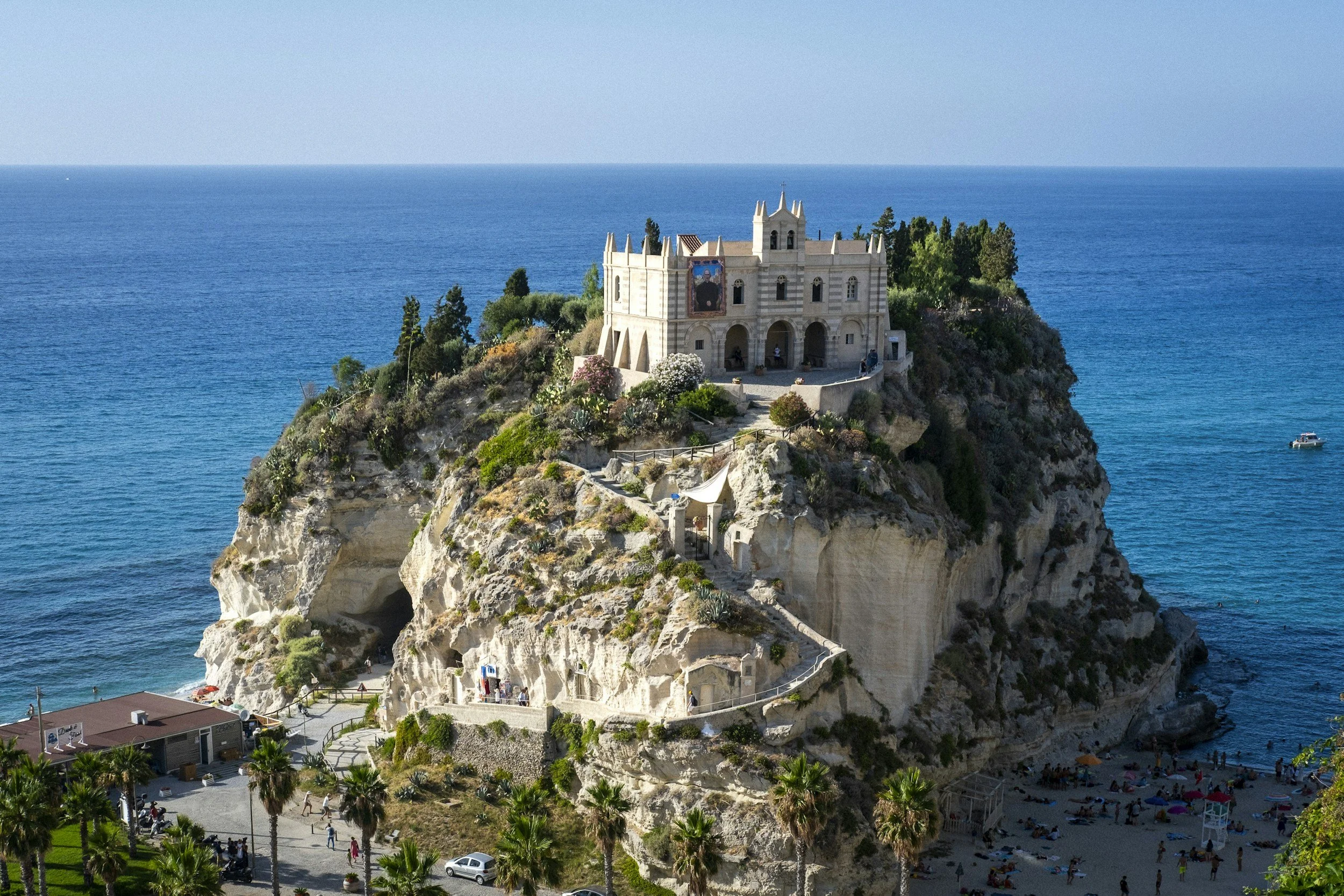 A chapel on a rocky outcrop overlooking the ocean, with a beach and palm trees below and boats on the water.