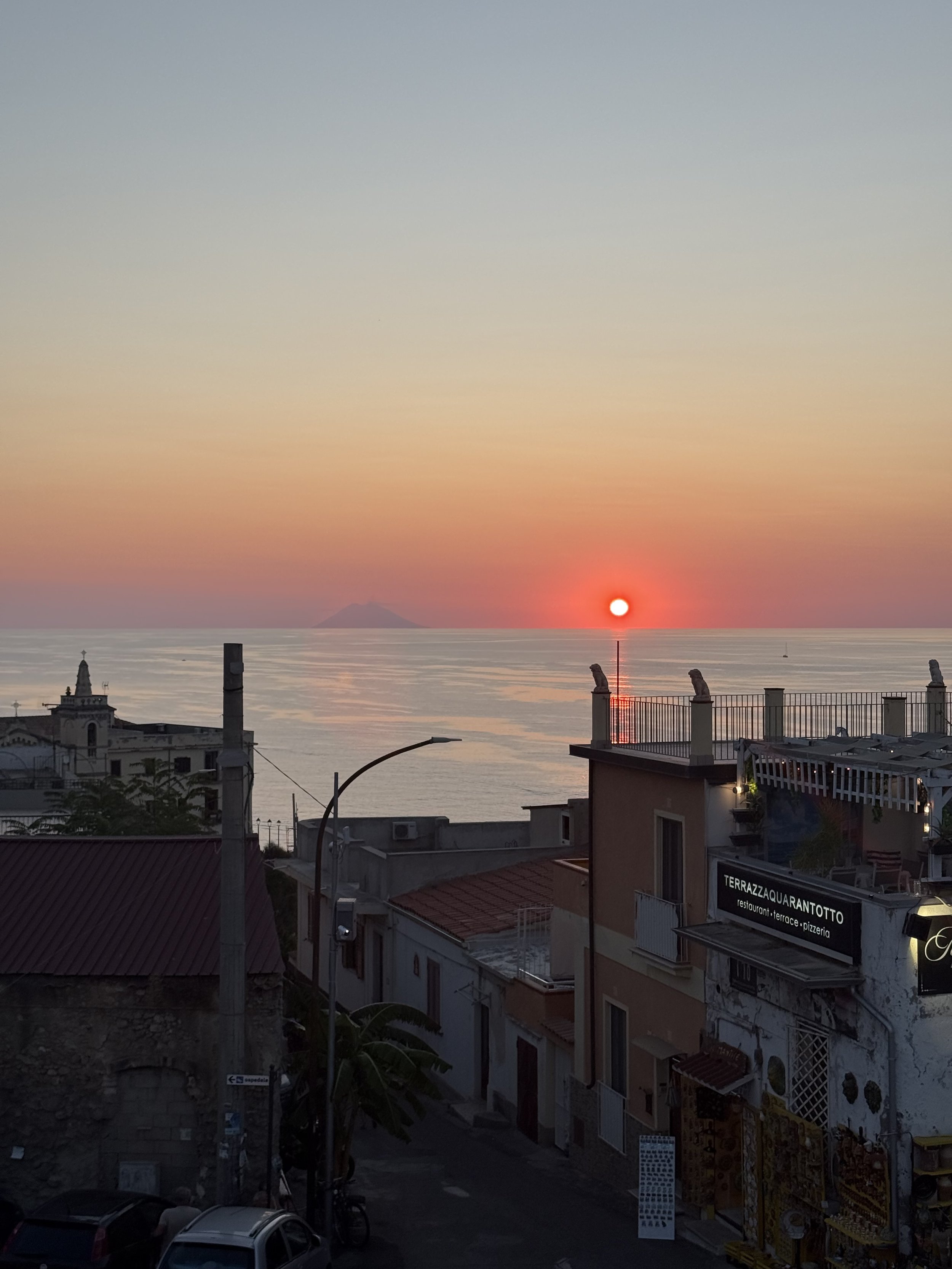 Sun setting over a coastal town with buildings and a view of the sea and distant mountains, warm colors in the sky
