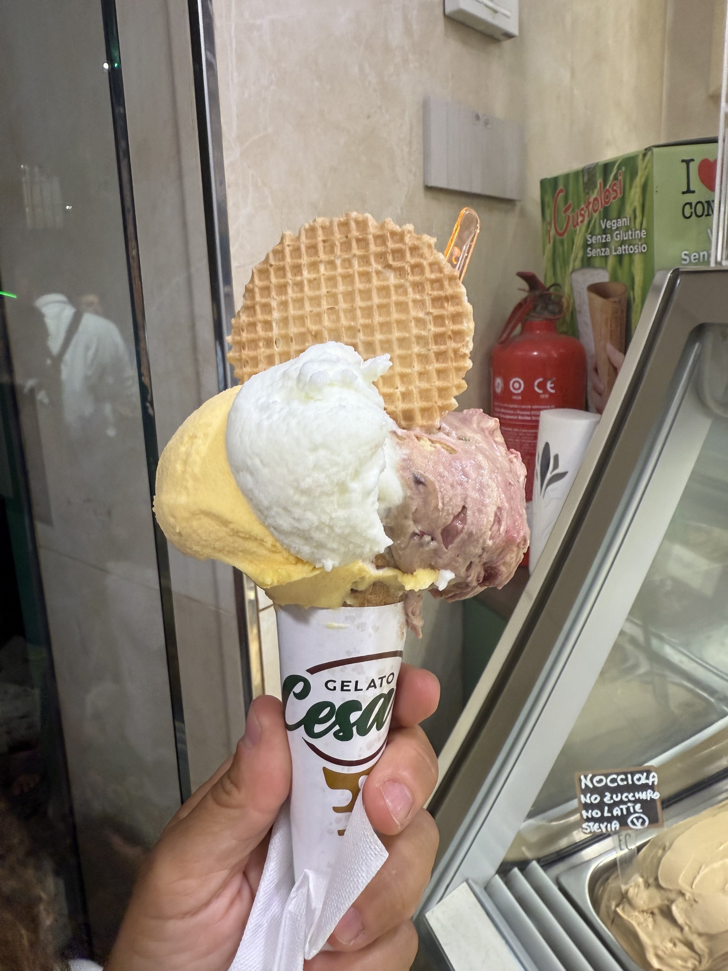 A person holding a cone of gelato with two waffle cookies on top, in front of an ice cream display case and a sign with written notes.