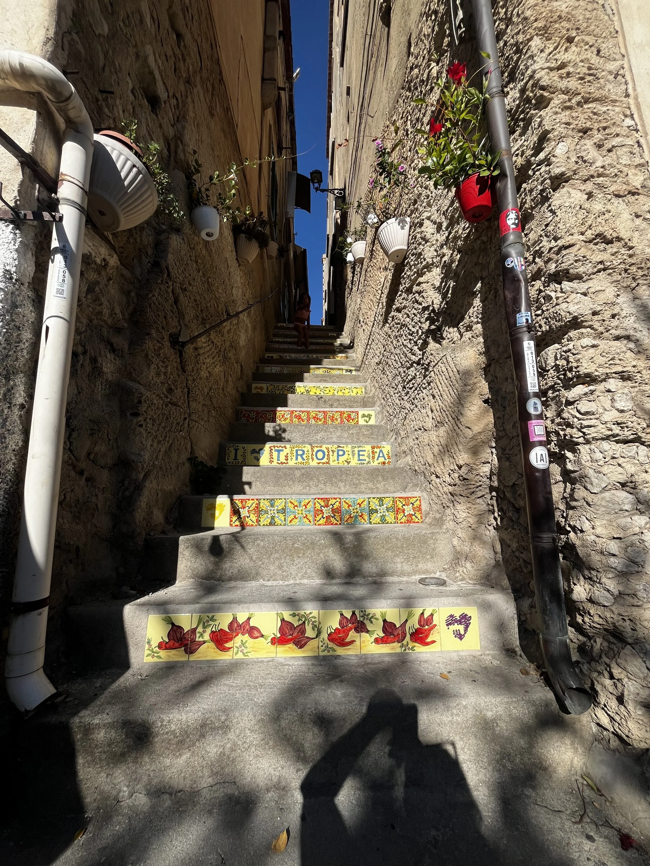 Street stairs with decorative tiles spelling out 'I ♥ TROPÉA', surrounded by flower pots, stone walls, and drain pipes.