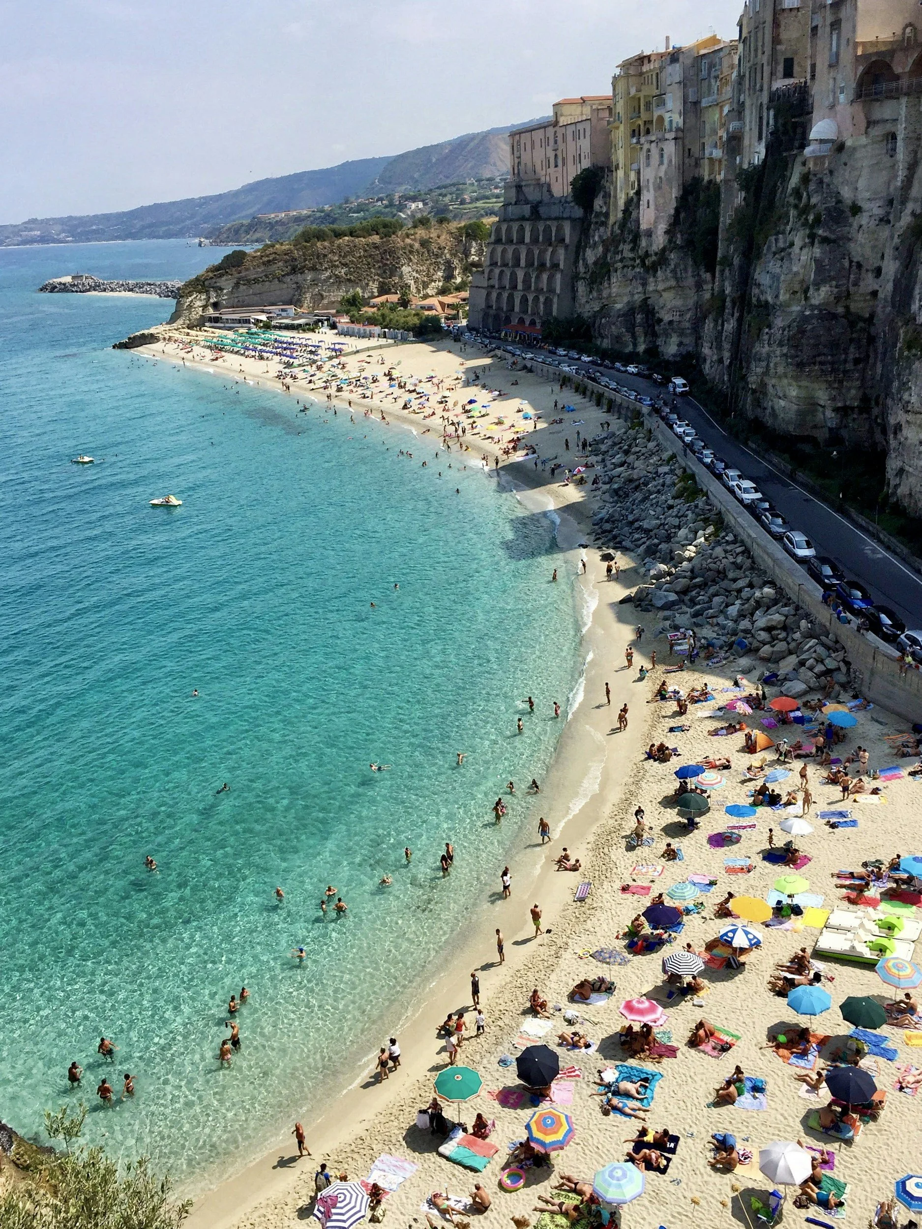 A crowded beach with colorful umbrellas and towels on the sand, people swimming and relaxing. The clear turquoise water extends to rocky breakwaters and a cliffside with buildings above.