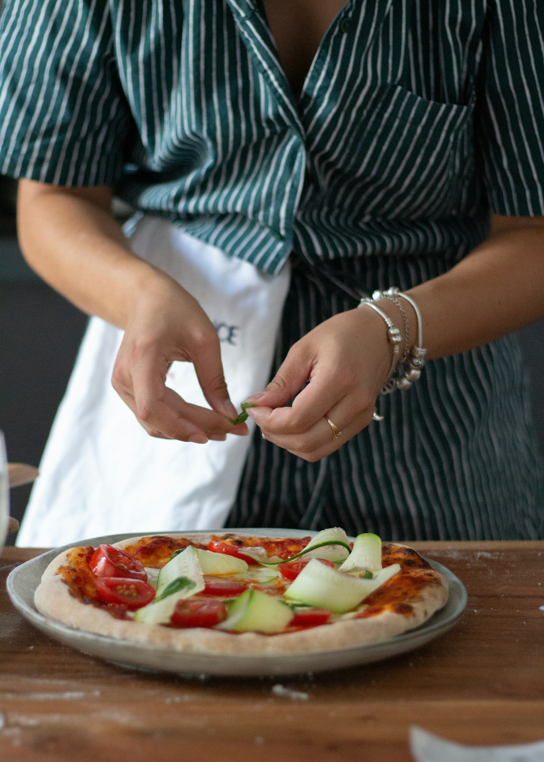 A person wearing a green and white striped shirt is preparing to add toppings to a pizza with sliced tomatoes and cucumbers on a wooden table in front of them.